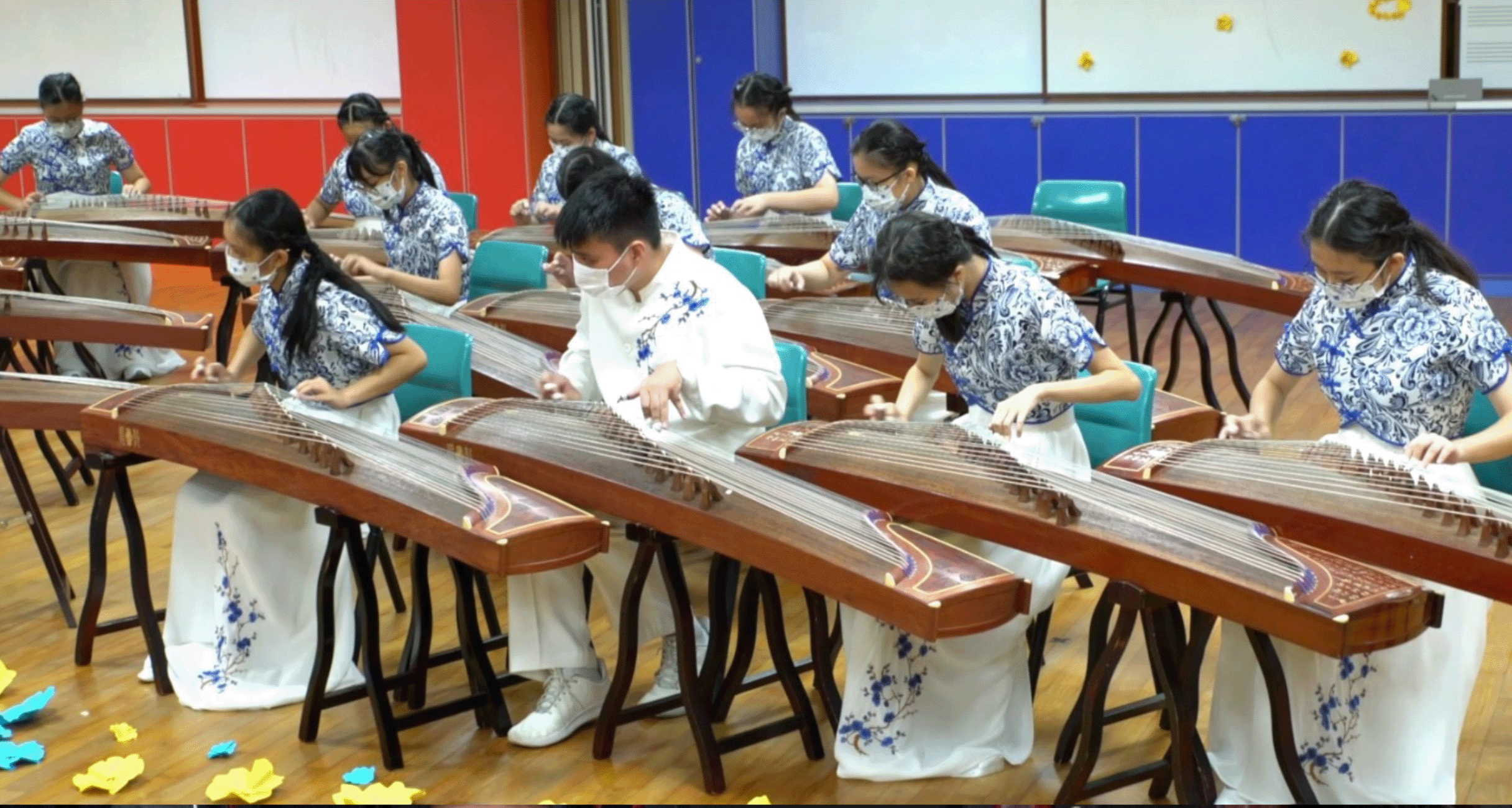 Several masked students in traditional Chinese attire play guzheng instruments in a classroom setting.
