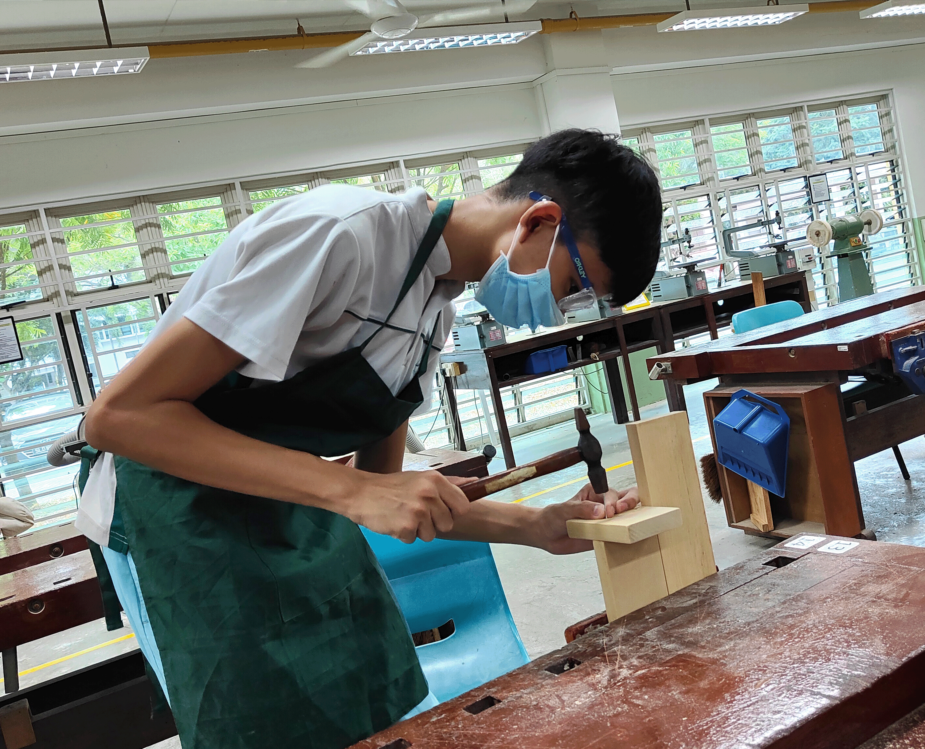Student in apron and mask chisels a wood joint at a workbench in a bright workshop.