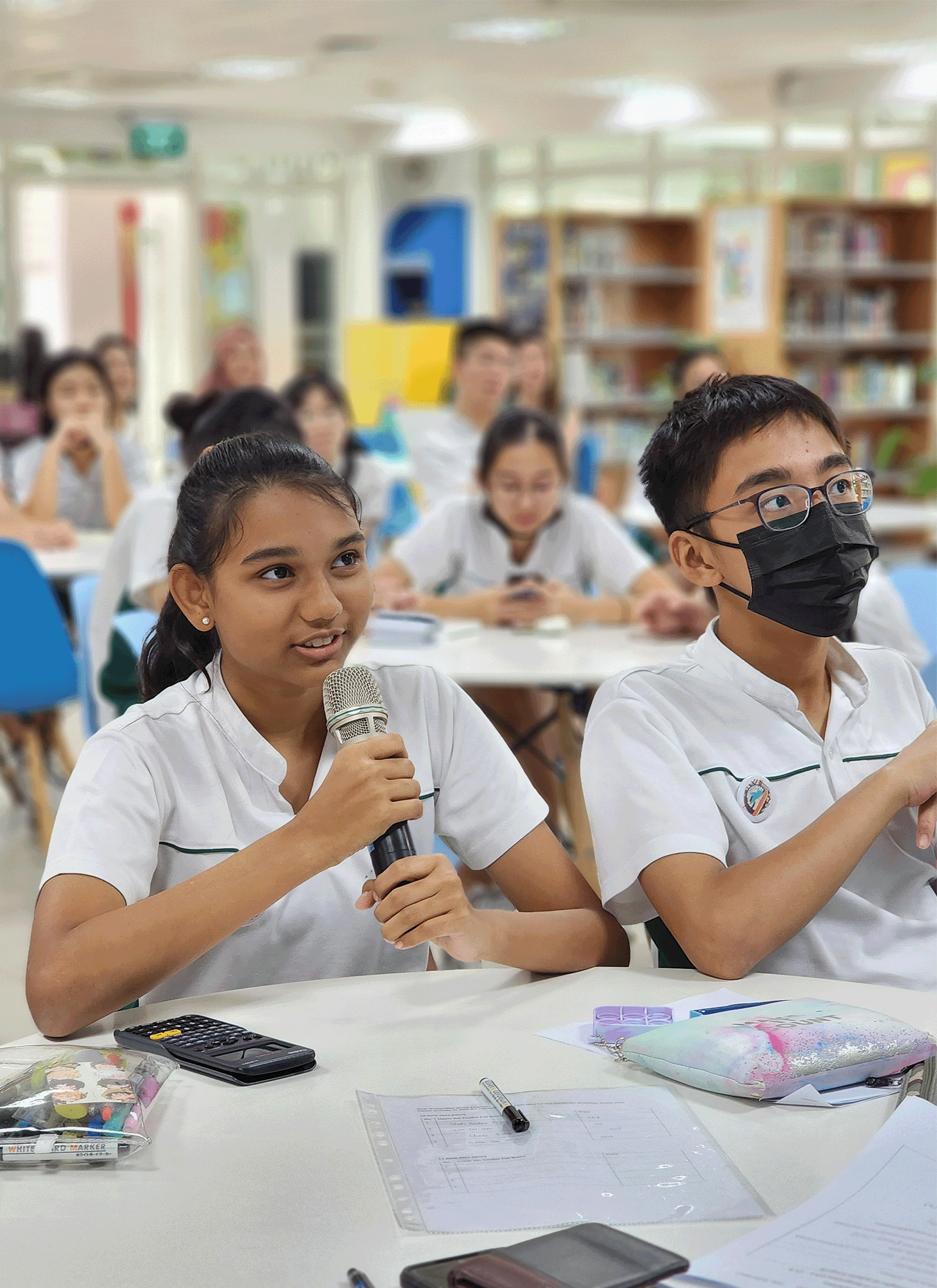 A girl in white uniform holds a microphone speaking, seated next to a masked boy with glasses in a school library.