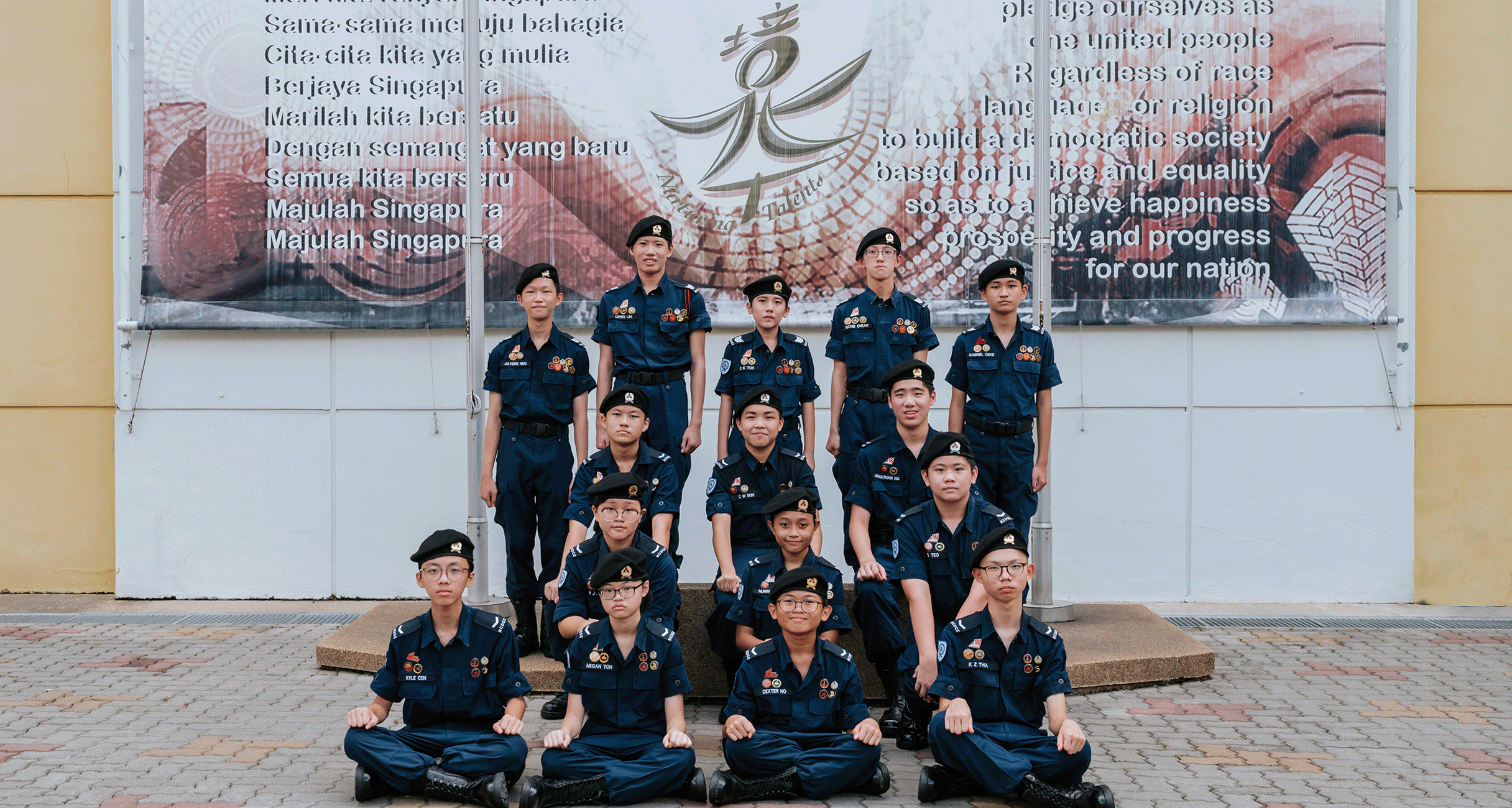 Group of young students in dark blue uniforms and black berets posing in three rows before a Singapore pledge banner.