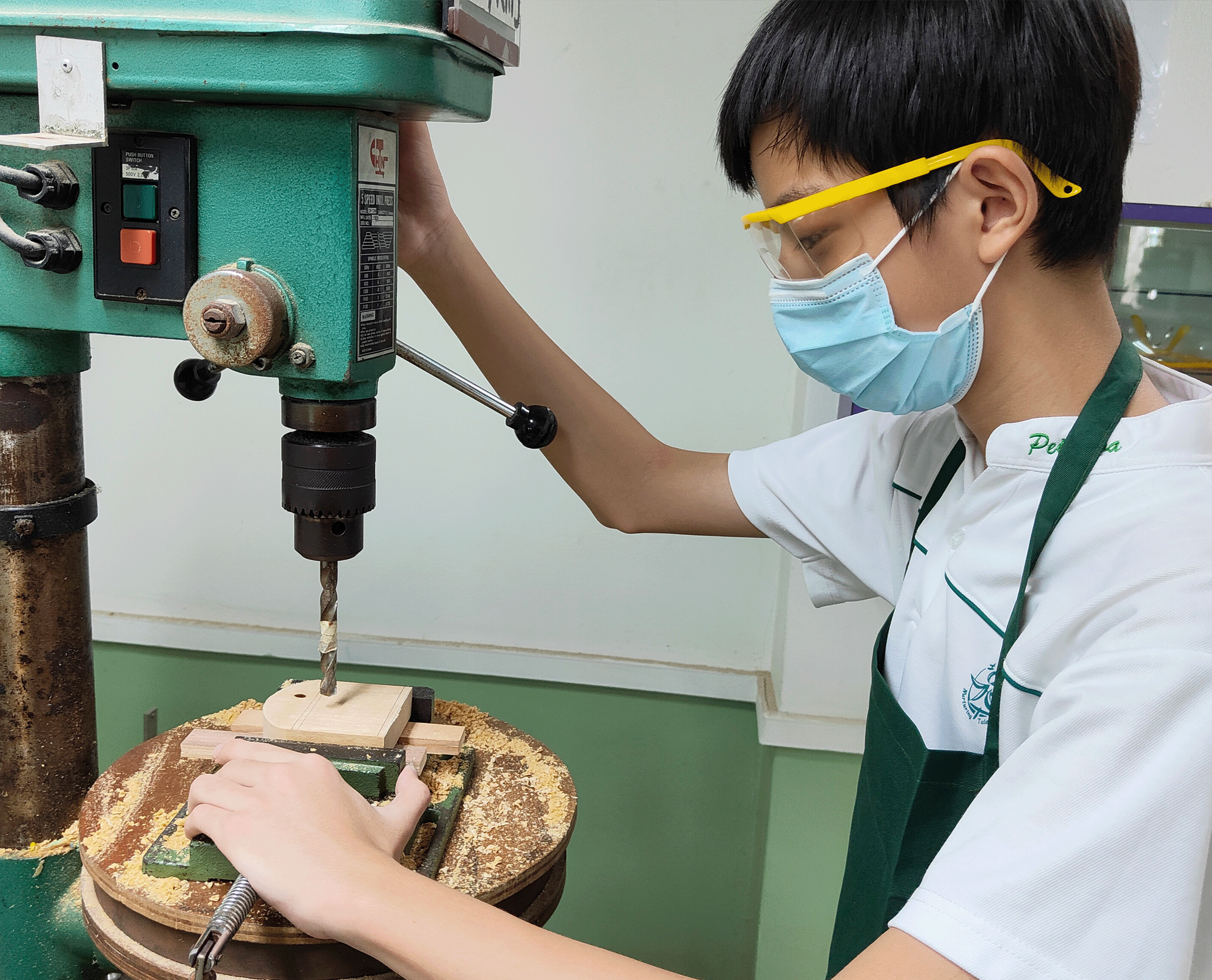 Student wearing goggles and mask operates a drill press, drilling a wood block clamped to the table.