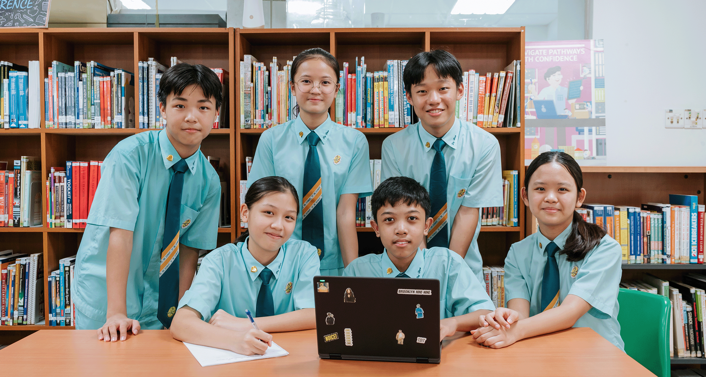 Six students in teal uniforms pose together in a school library with a sticker-covered laptop on the table.