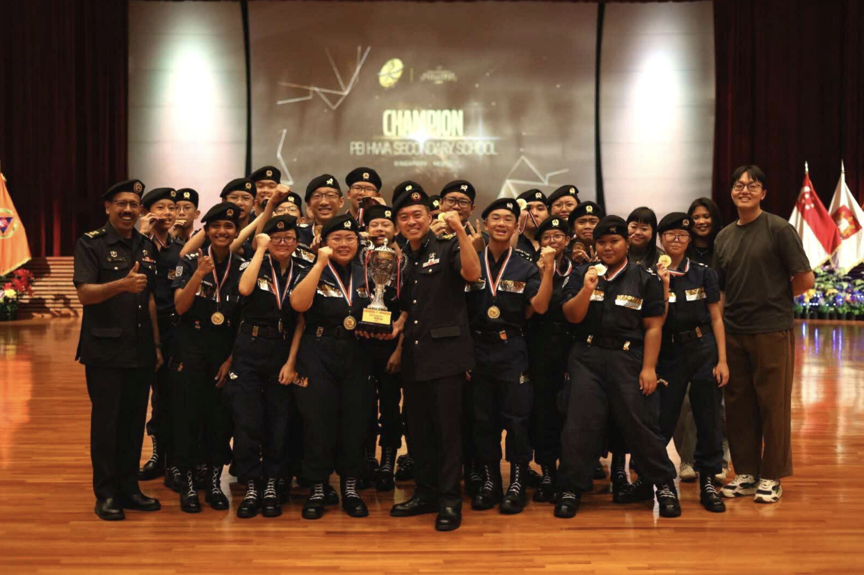 Group of uniformed students with gold medals and a trophy posing on stage under a "Champion" banner.