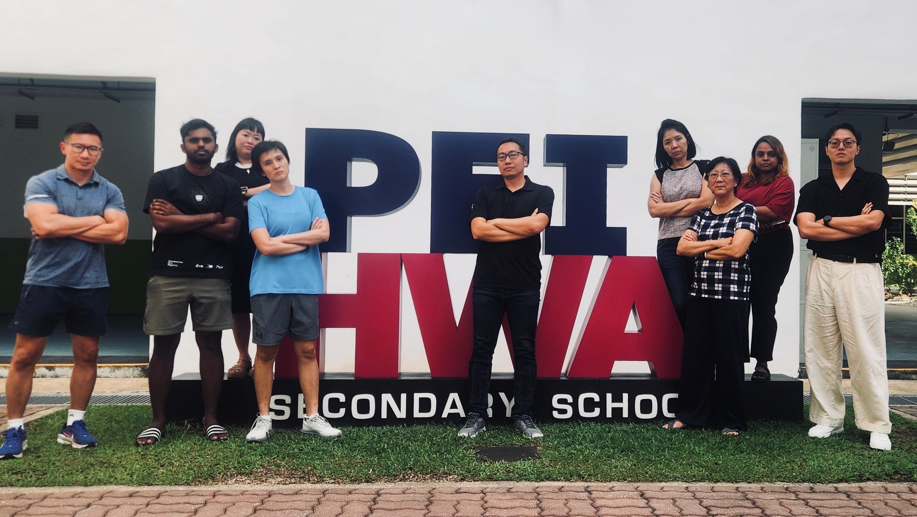 Group of about 13 people posing with arms crossed pose in front of a large Pei Hwa Secondary School sign outdoors.