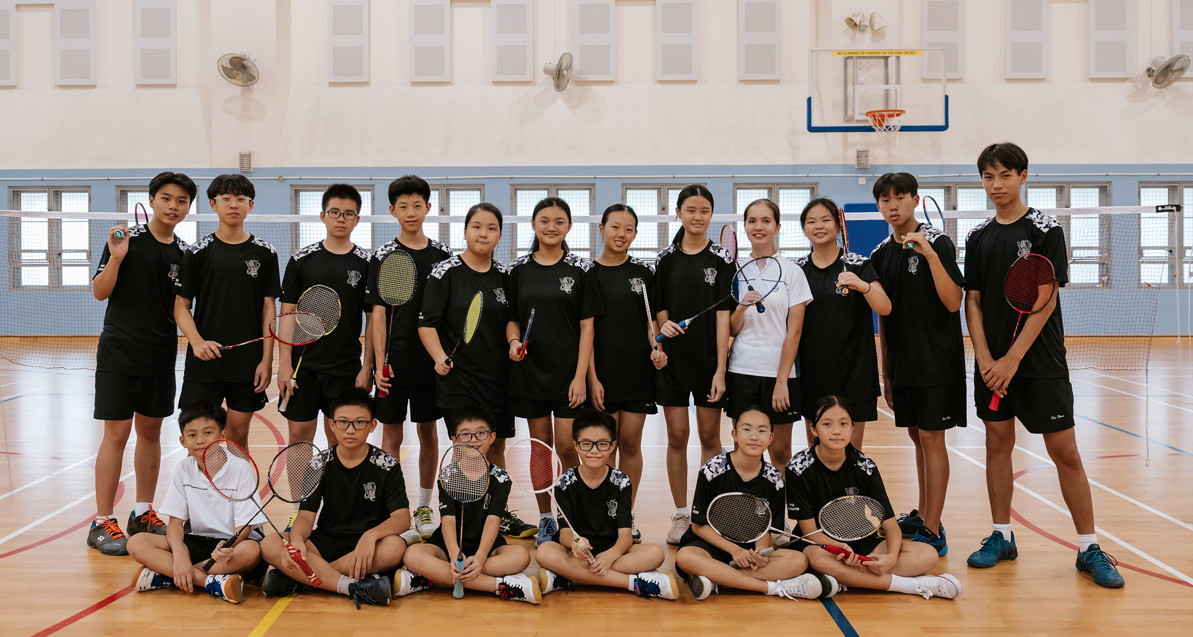 Group of badminton players in black uniforms posing in a gym, holding rackets; some seated in front.