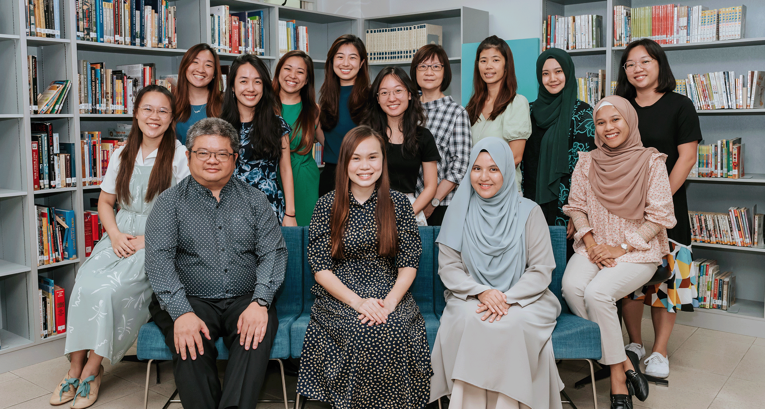 A group of about 15 smiling people posing together in a library with bookshelves in the background.