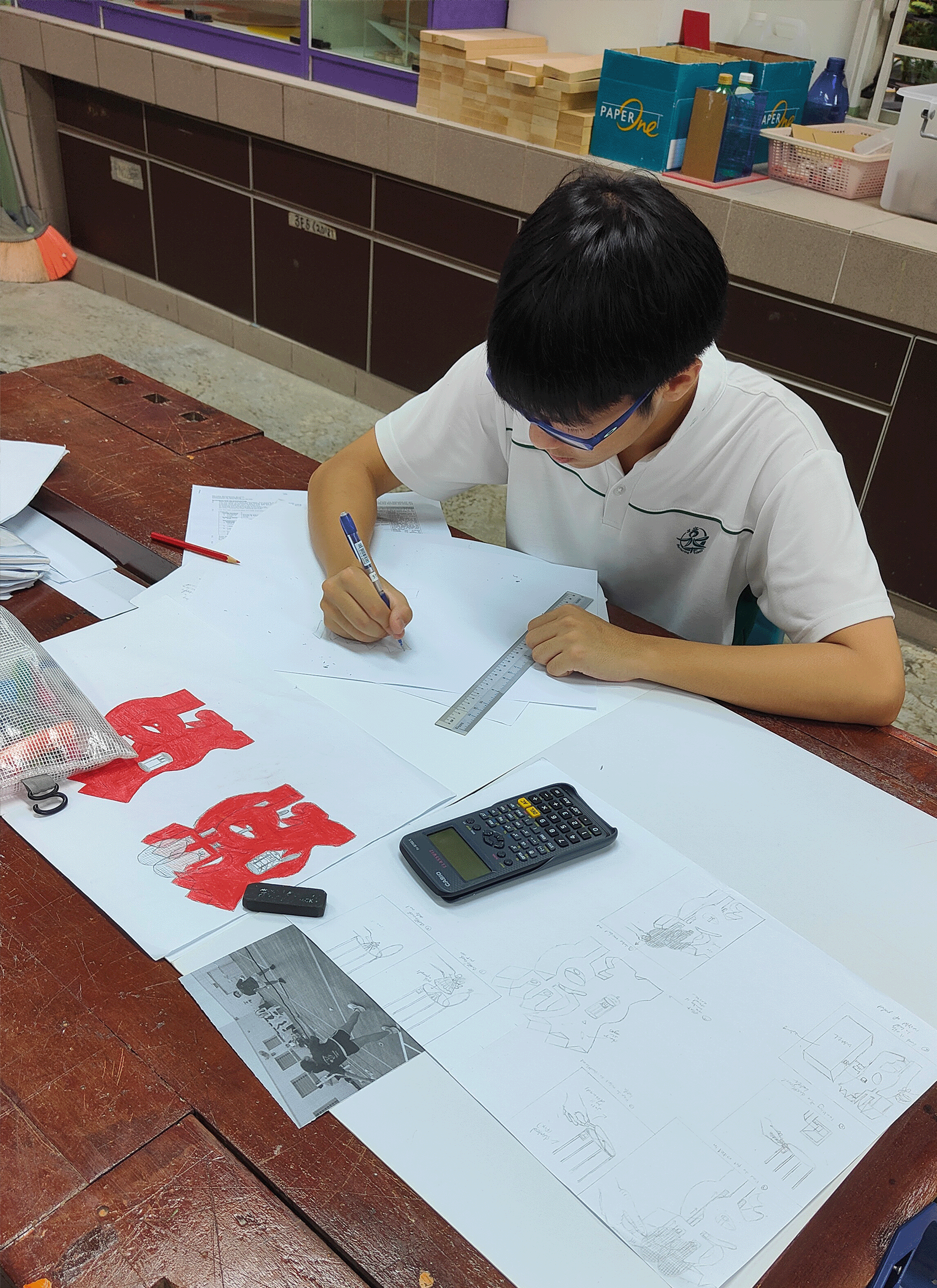 A young student wearing glasses draws on large paper sheets with a ruler and calculator on a wooden desk.