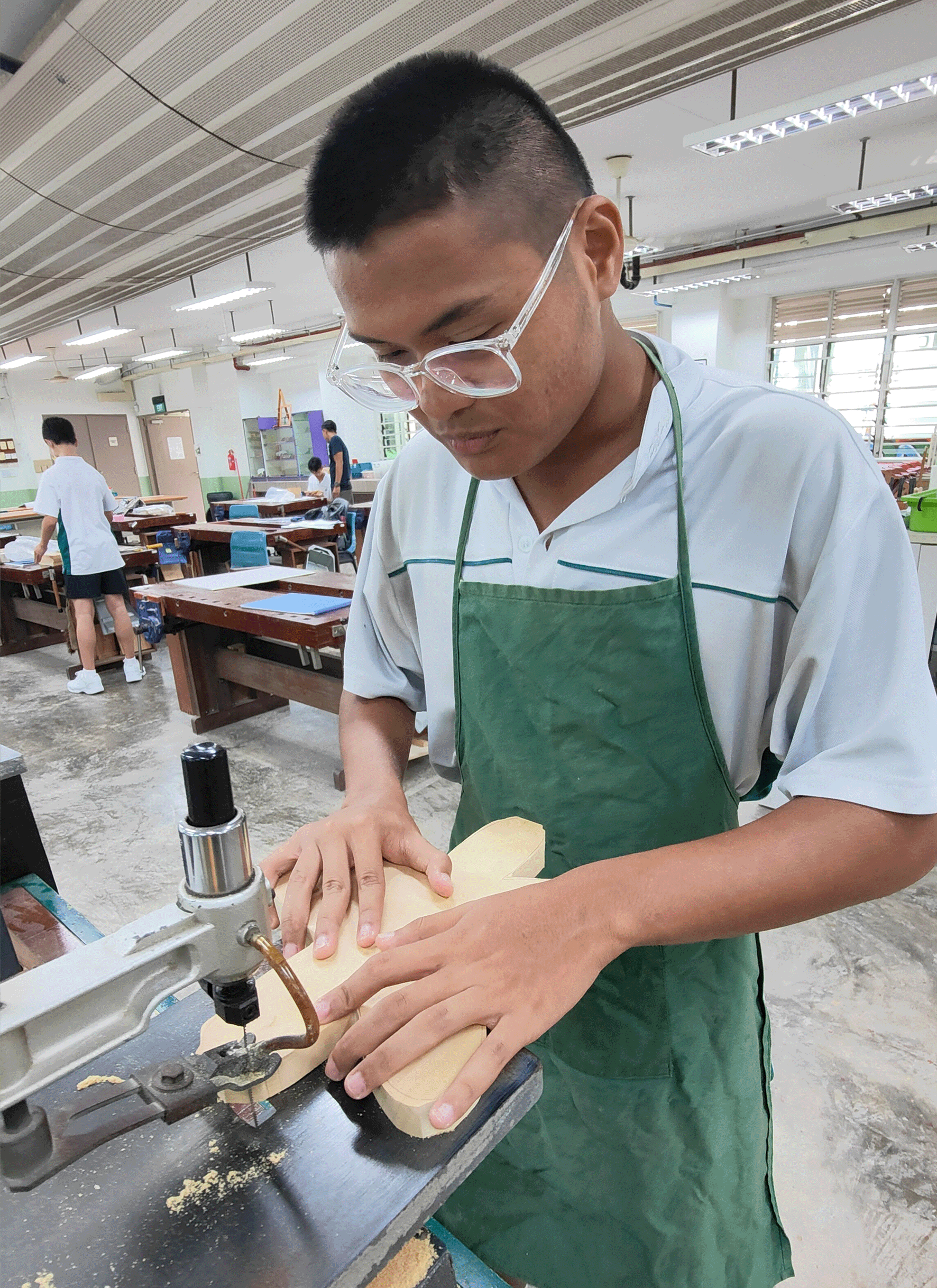 Young man wearing safety goggles and green apron cuts a wood block using a band saw in a workshop classroom.