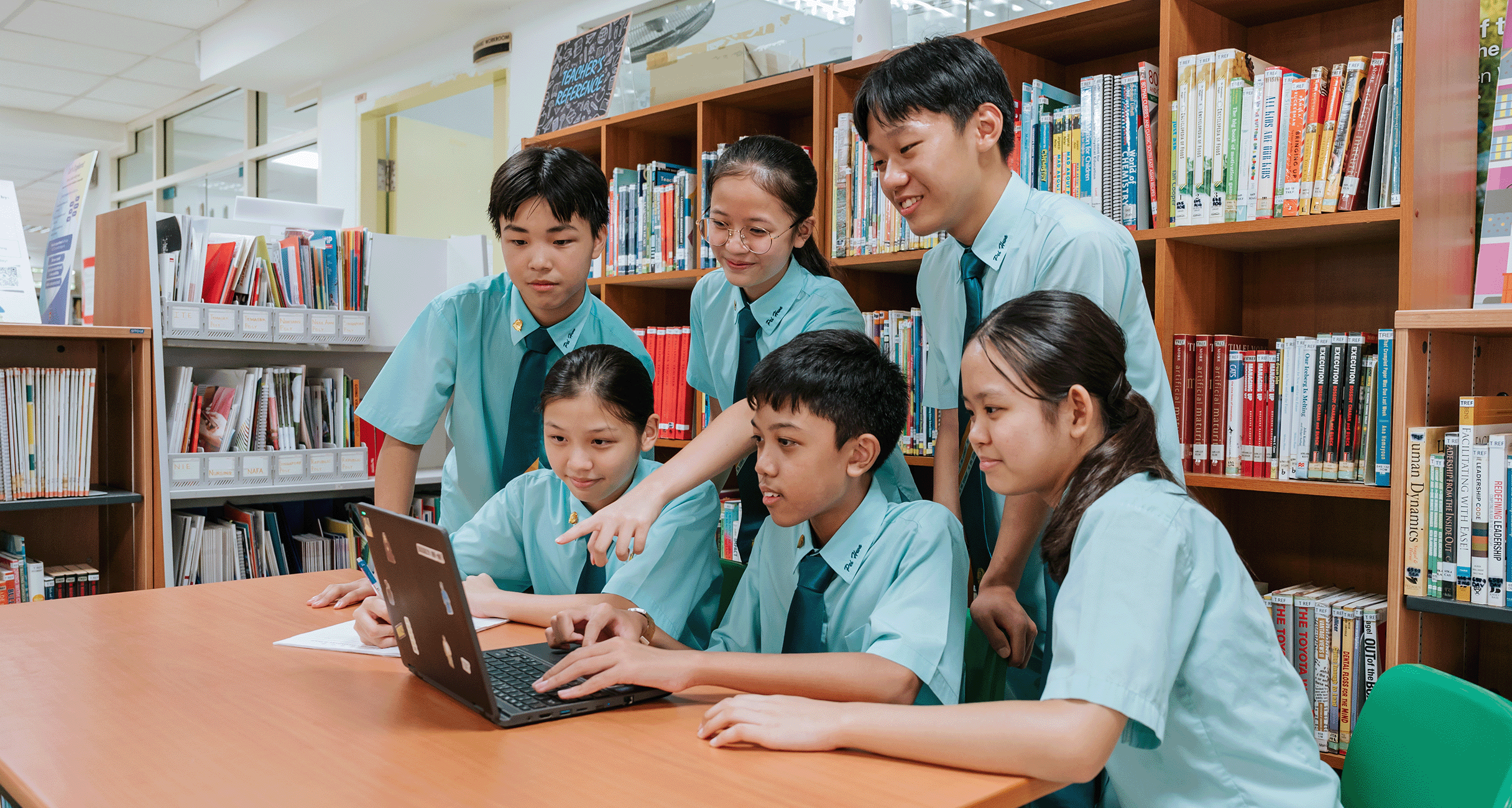 Six students in teal uniforms gather around a laptop at a library table, surrounded by bookshelves.