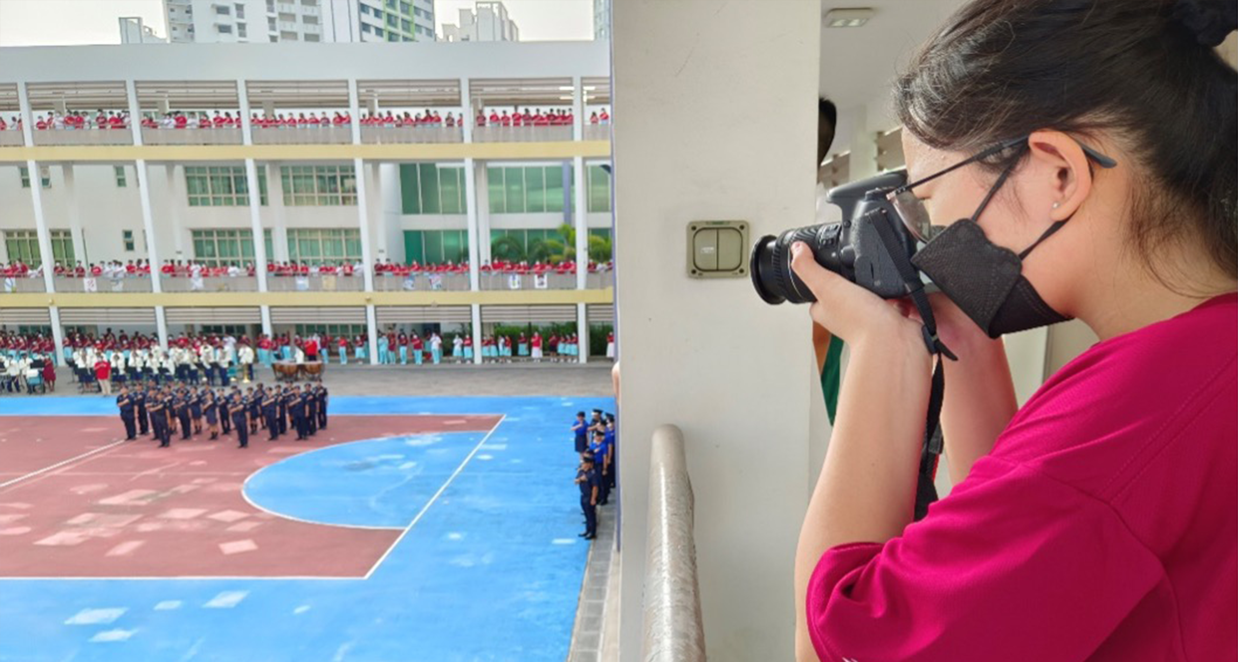 A masked woman in pink photographs a school assembly with uniformed students on a blue courtyard below.