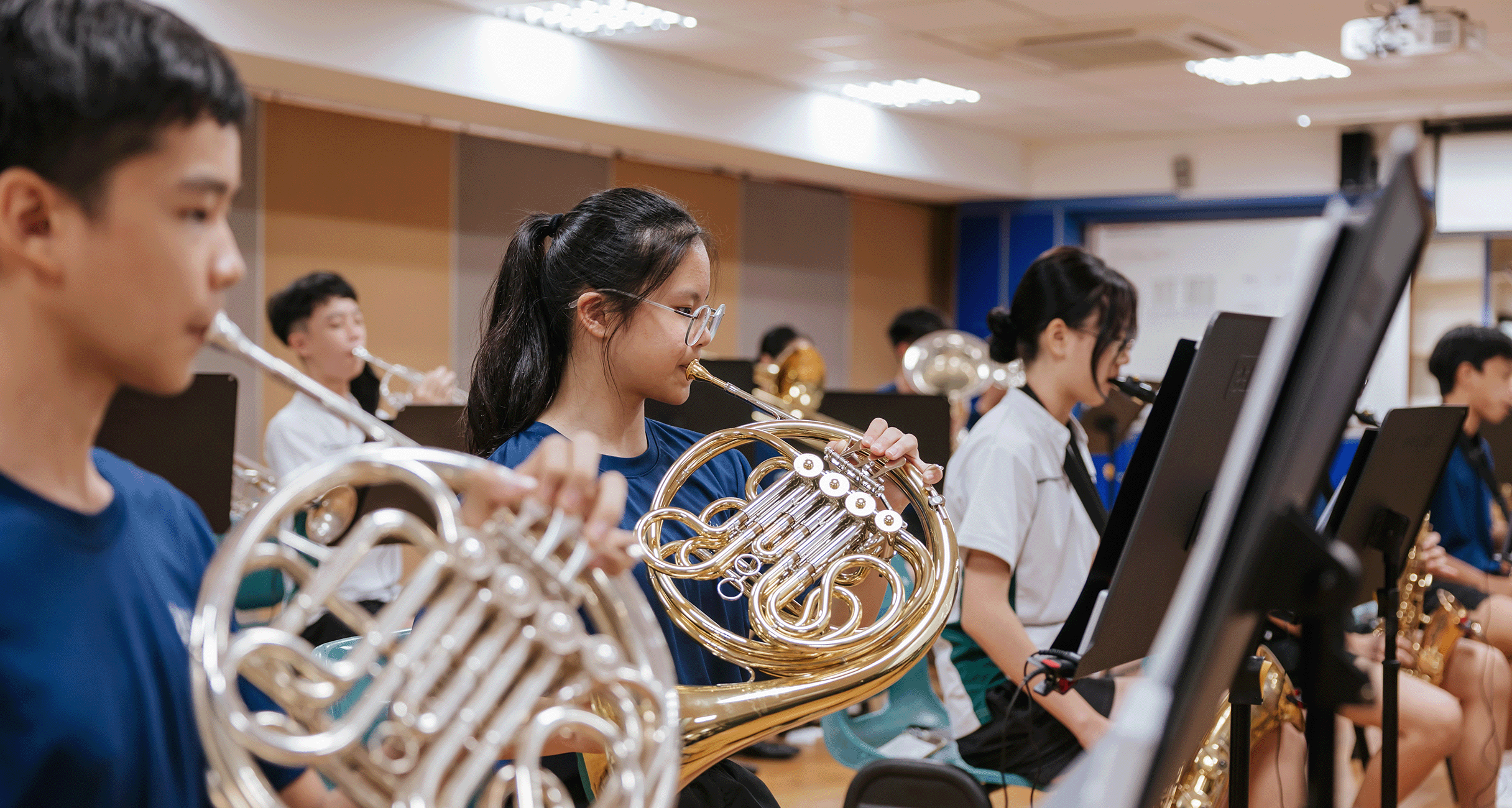 Students playing French horns and brass instruments during a band rehearsal in a classroom.