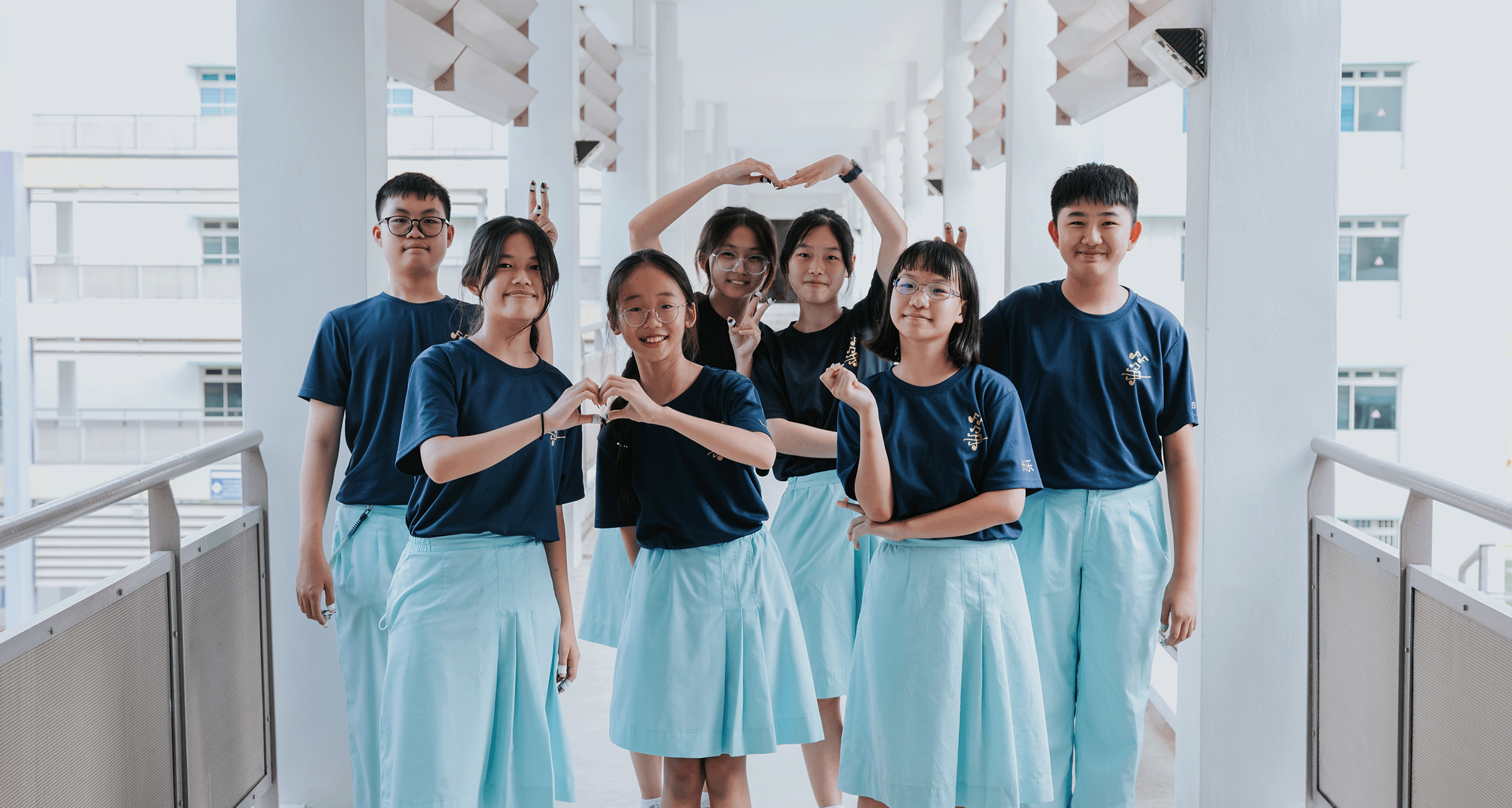 Seven students in CCA shirts and school uniform bottoms pose on a school corridor