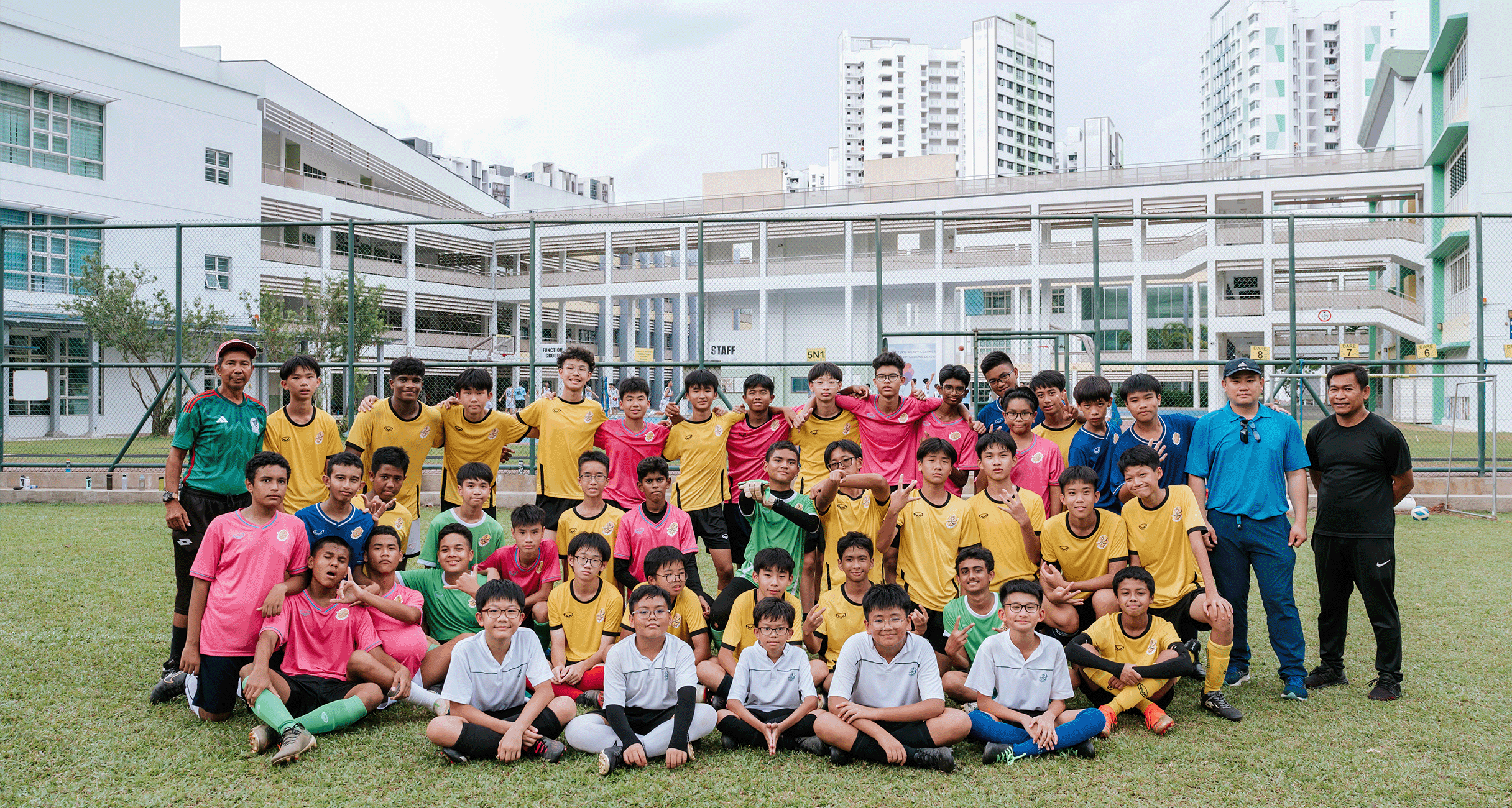 A large group of young male football players in yellow, pink, and white jerseys posing on a school field.