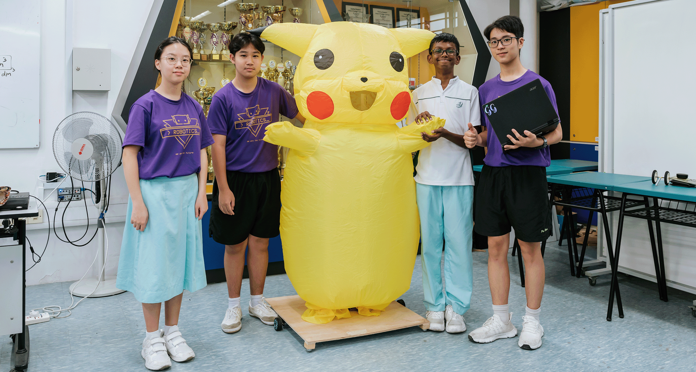 Four students in purple robotics shirts stand beside a large inflatable Pikachu costume in a trophy-lined classroom.
