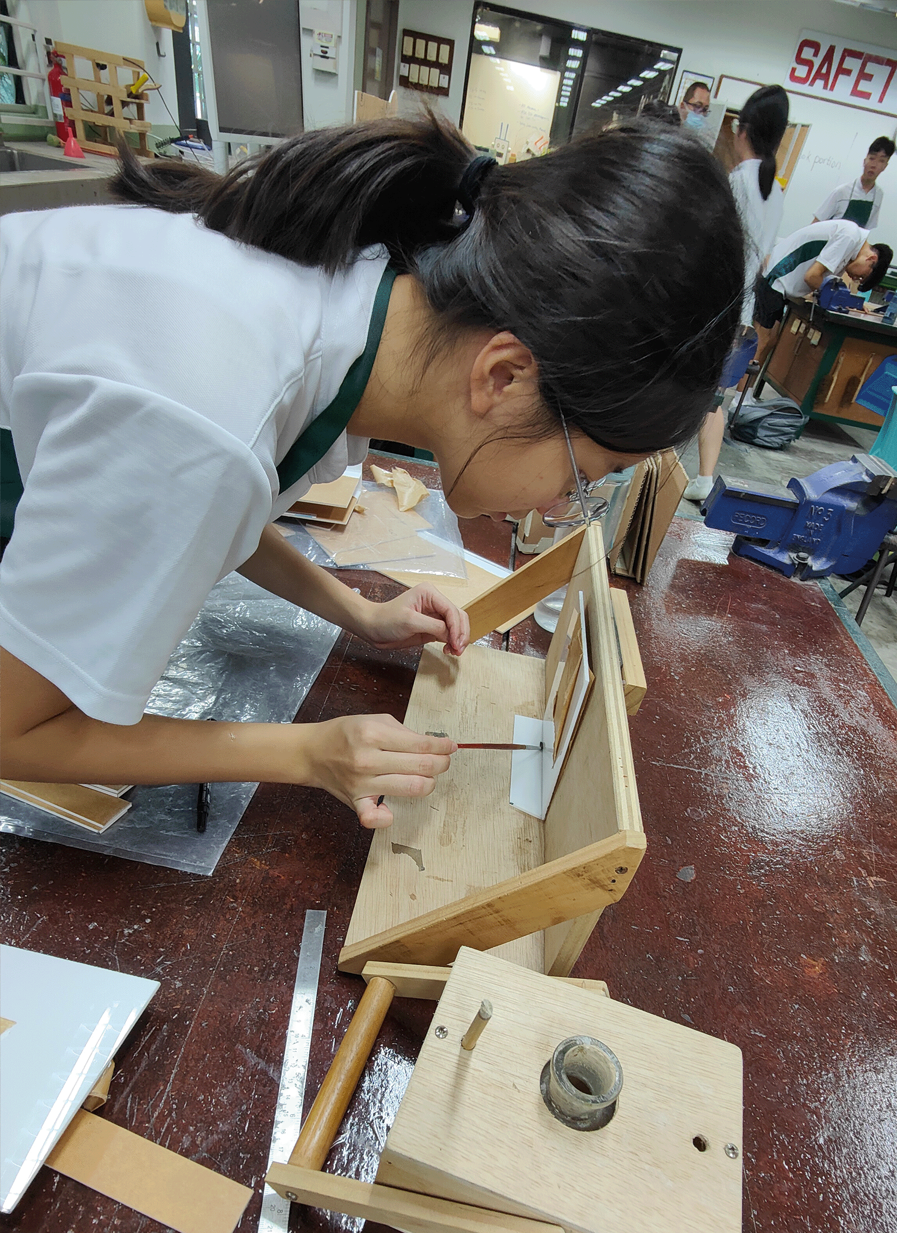A young woman in a white shirt leans over a workbench, carefully assembling a wooden structure in a workshop classroom.