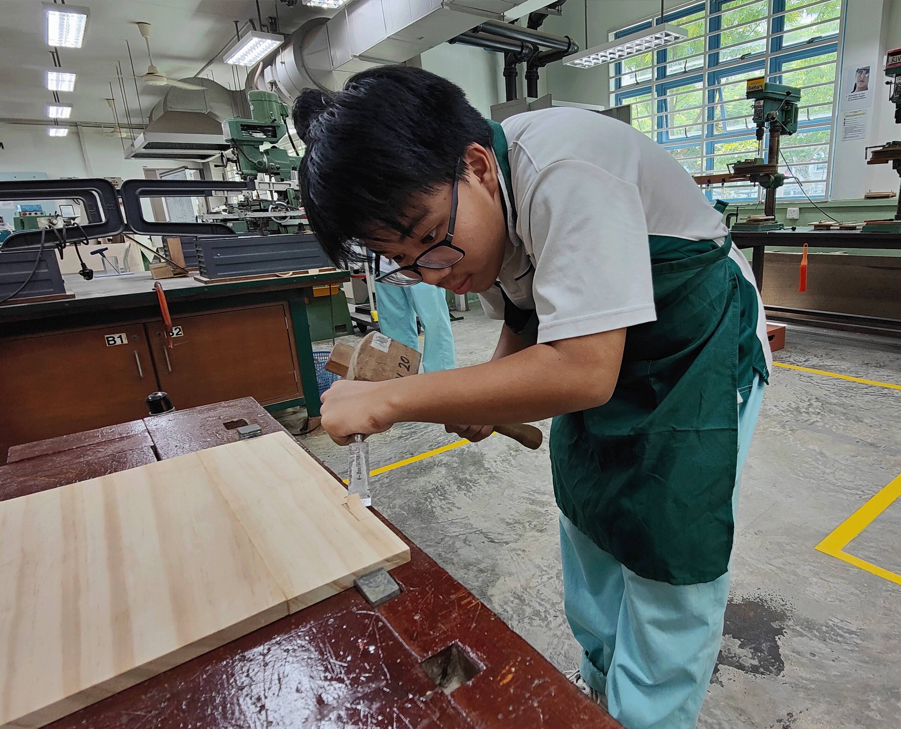 Student in a green apron chisels a wood board on a bench in a workshop with machines and windows.