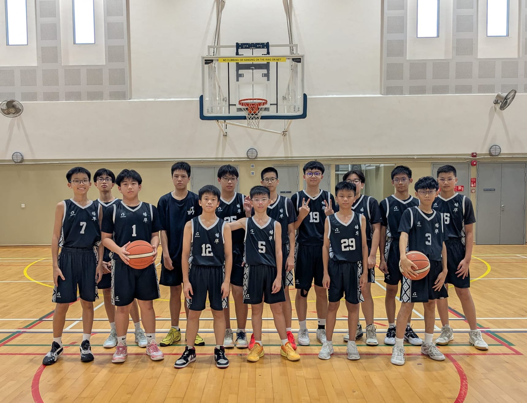 A youth basketball team of about 14 boys in black jerseys posing on an indoor court beneath a basketball hoop.