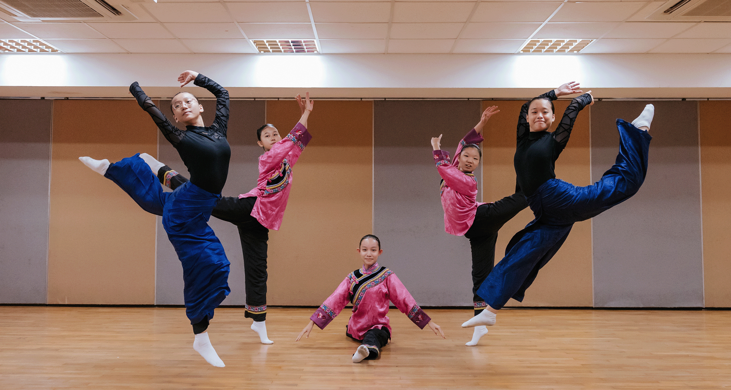 Five young dancers in traditional Chinese costumes pose in a studio, two leaping high with legs raised.