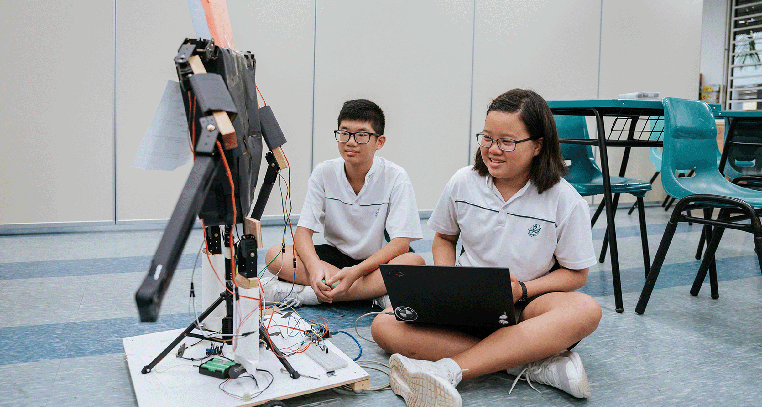 Two students in white uniforms sit on the floor with a laptop, working on a wired robotics project.