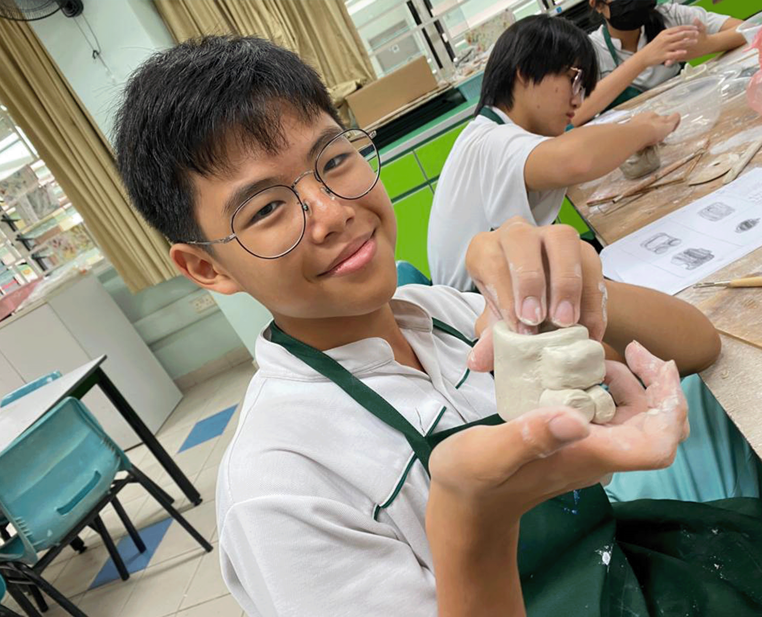Student in glasses smiles while holding a small clay sculpture at a classroom table with other students