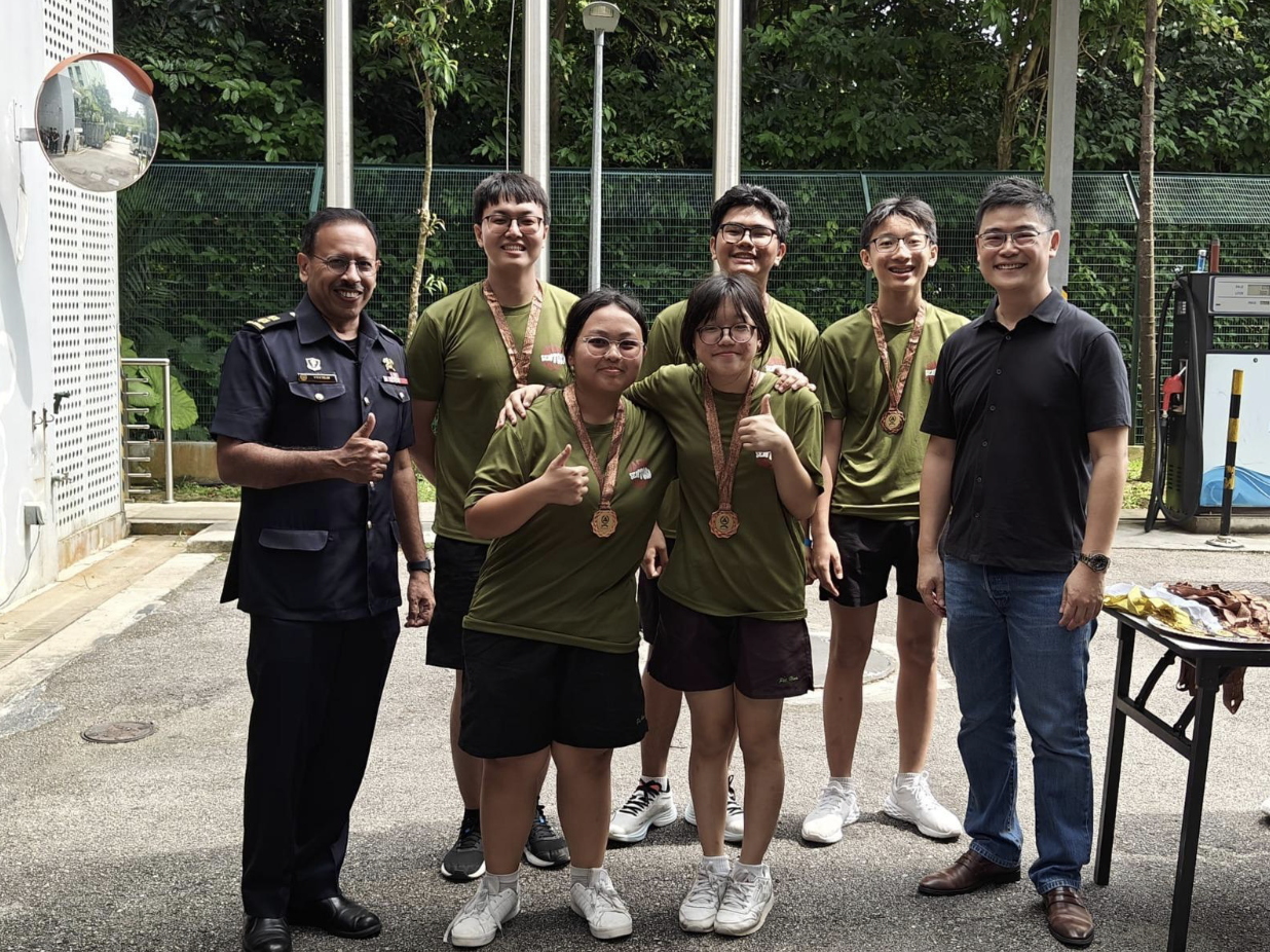 Seven people pose outdoors, five youths in olive shirts wearing medals, flanked by an officer and a man in black.