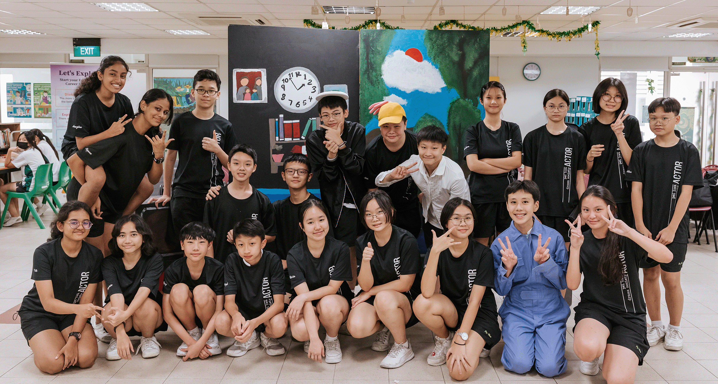 A large group of students in black "ACTOR" shirts pose together in front of a painted stage backdrop indoors.
