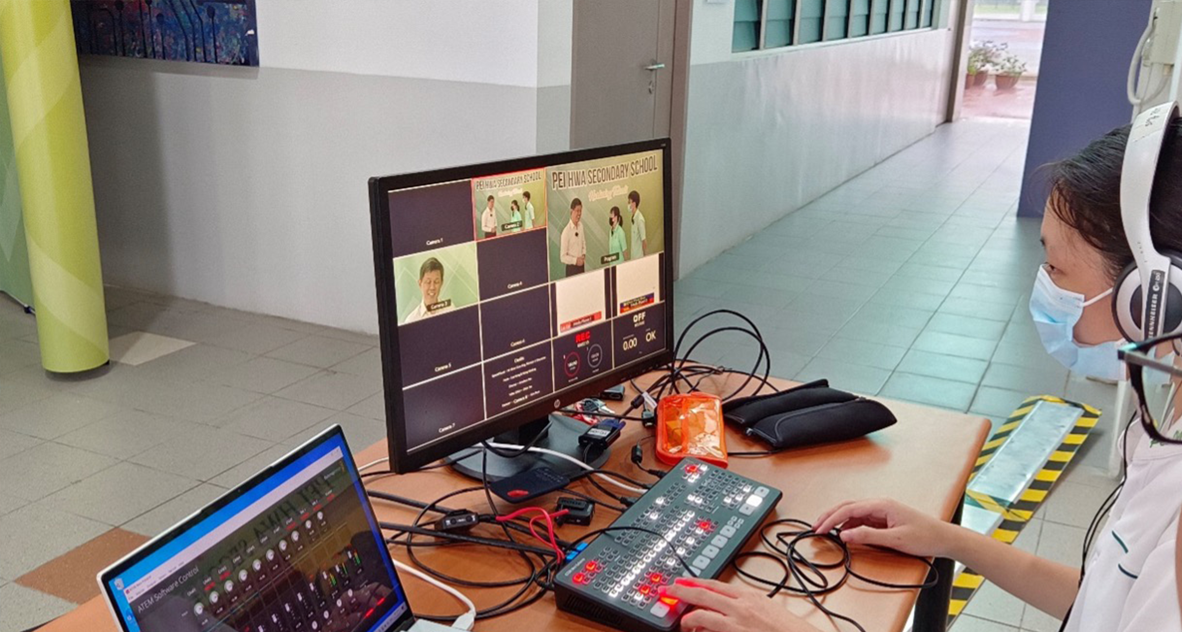Person wearing a mask and headphones operates a video switcher at a desk with a monitor showing a live school broadcast.