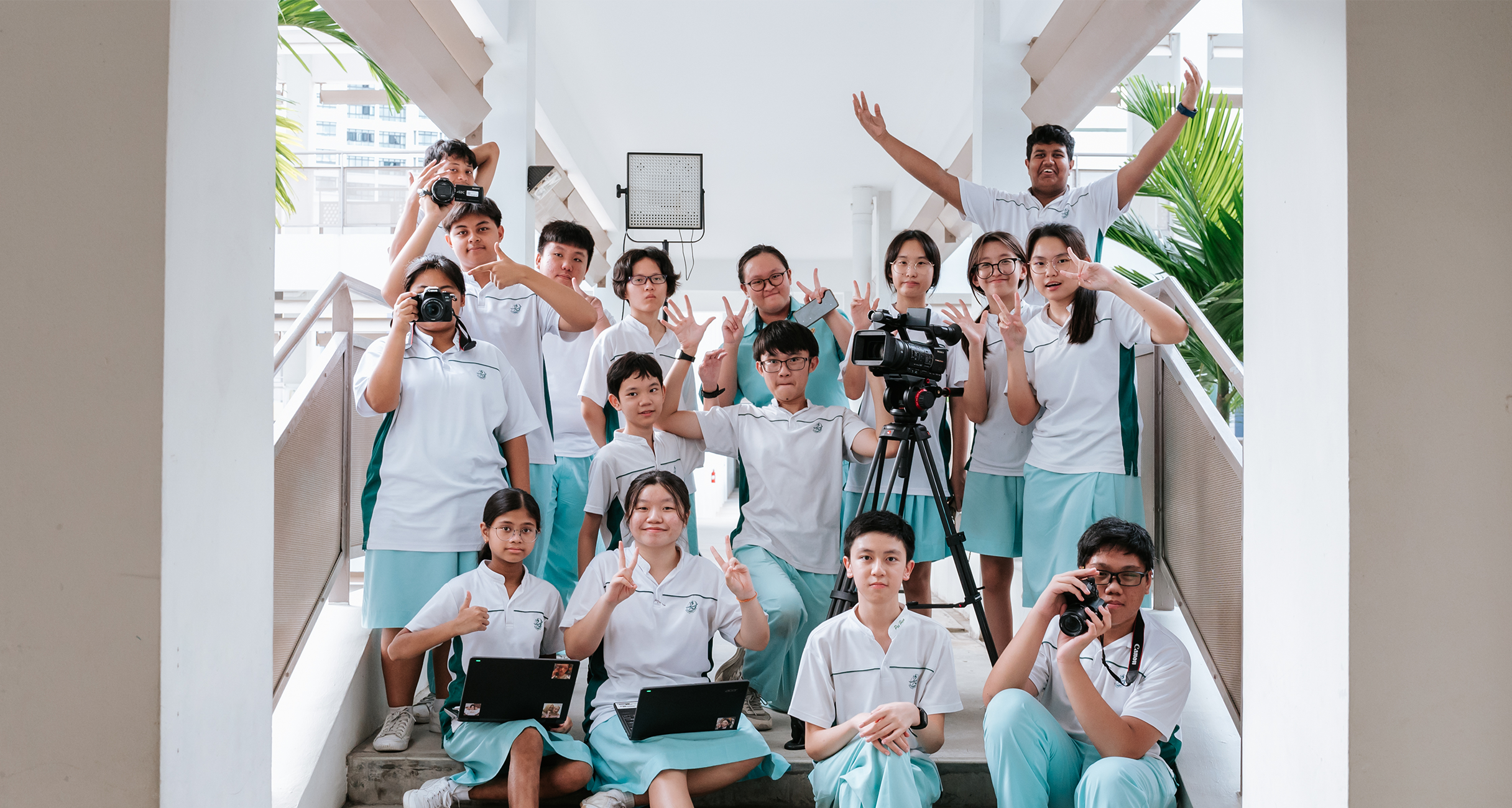 Group of students in white uniforms pose on stairs with cameras, tripod, and video light.