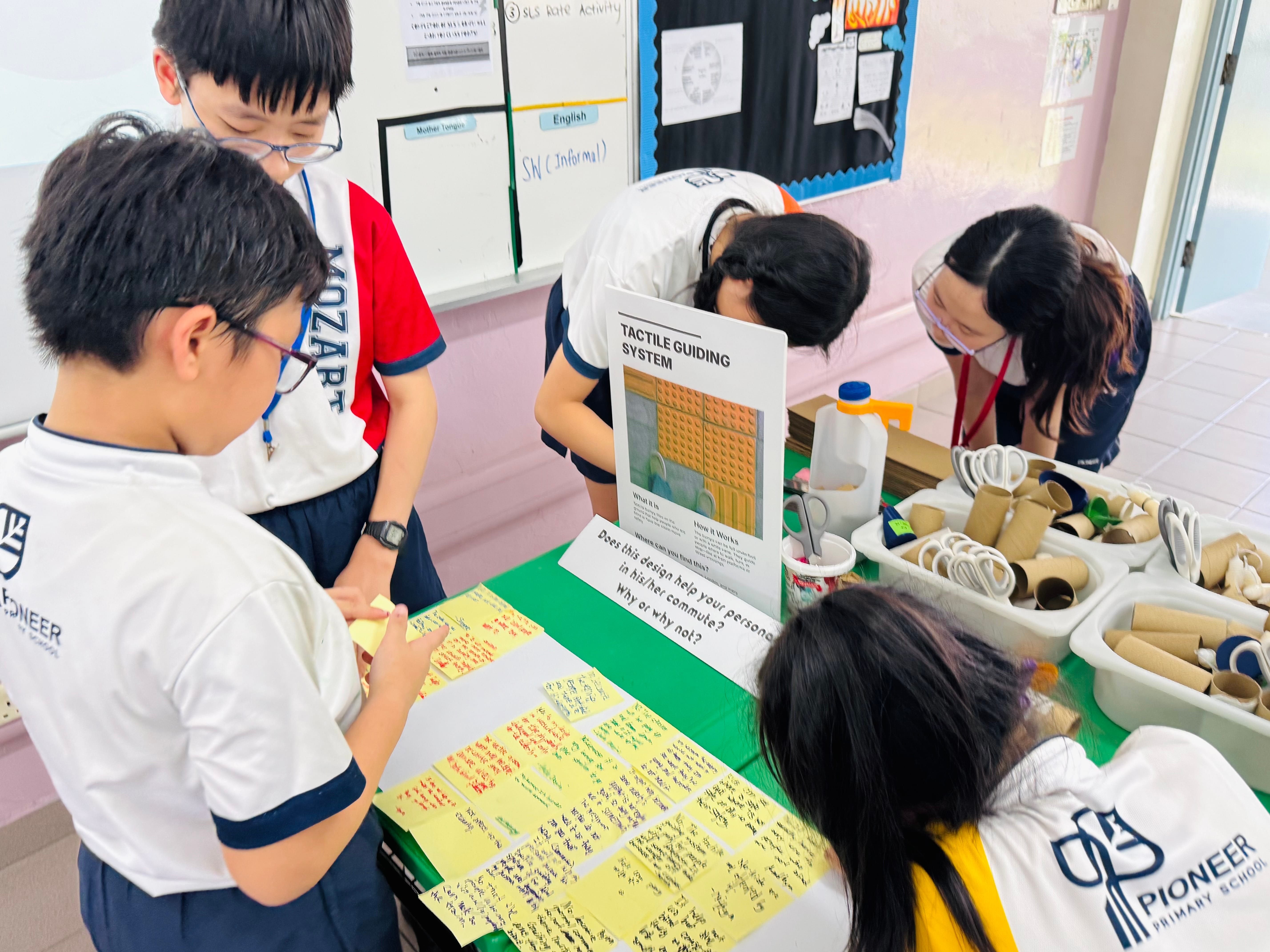 Students in school uniforms gather around a table with a Tactile Guiding System poster and sticky notes.
