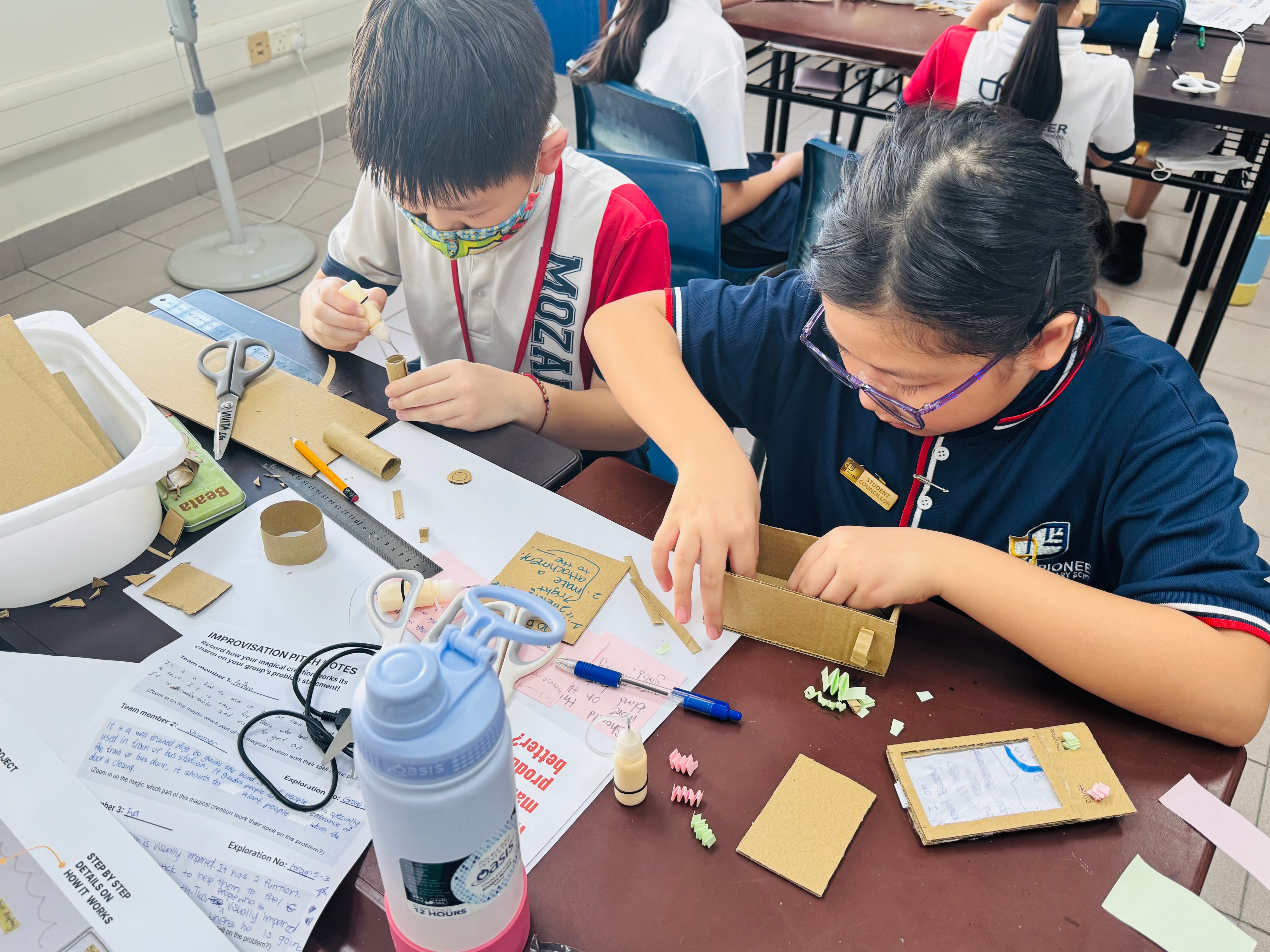 Two students at a desk build cardboard crafts with scissors, rulers, glue, and paper scraps.
