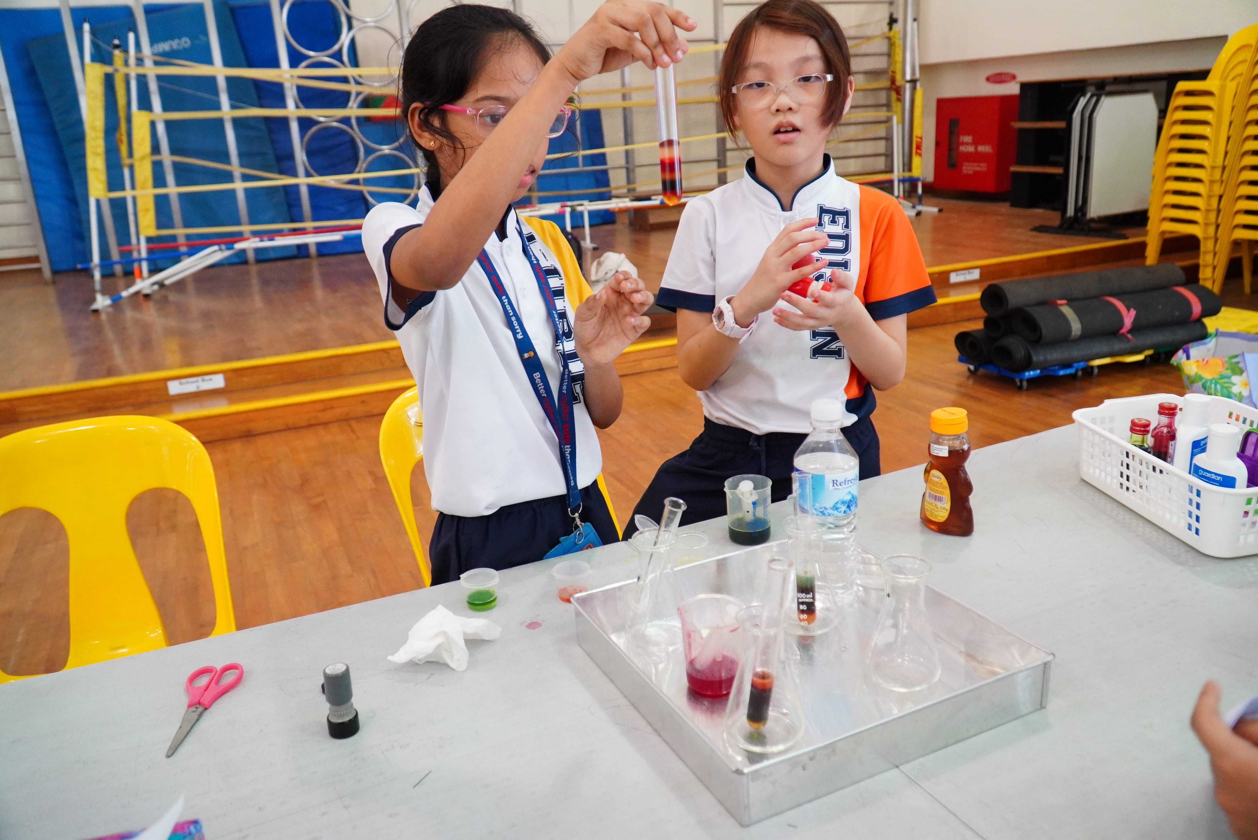 Two students in goggles do a science experiment, holding a test tube over a table of beakers and liquids.