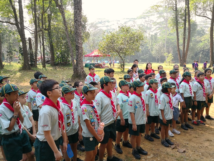 Group of scouts in uniform with red neckerchiefs standing in lines outdoors among tall trees.