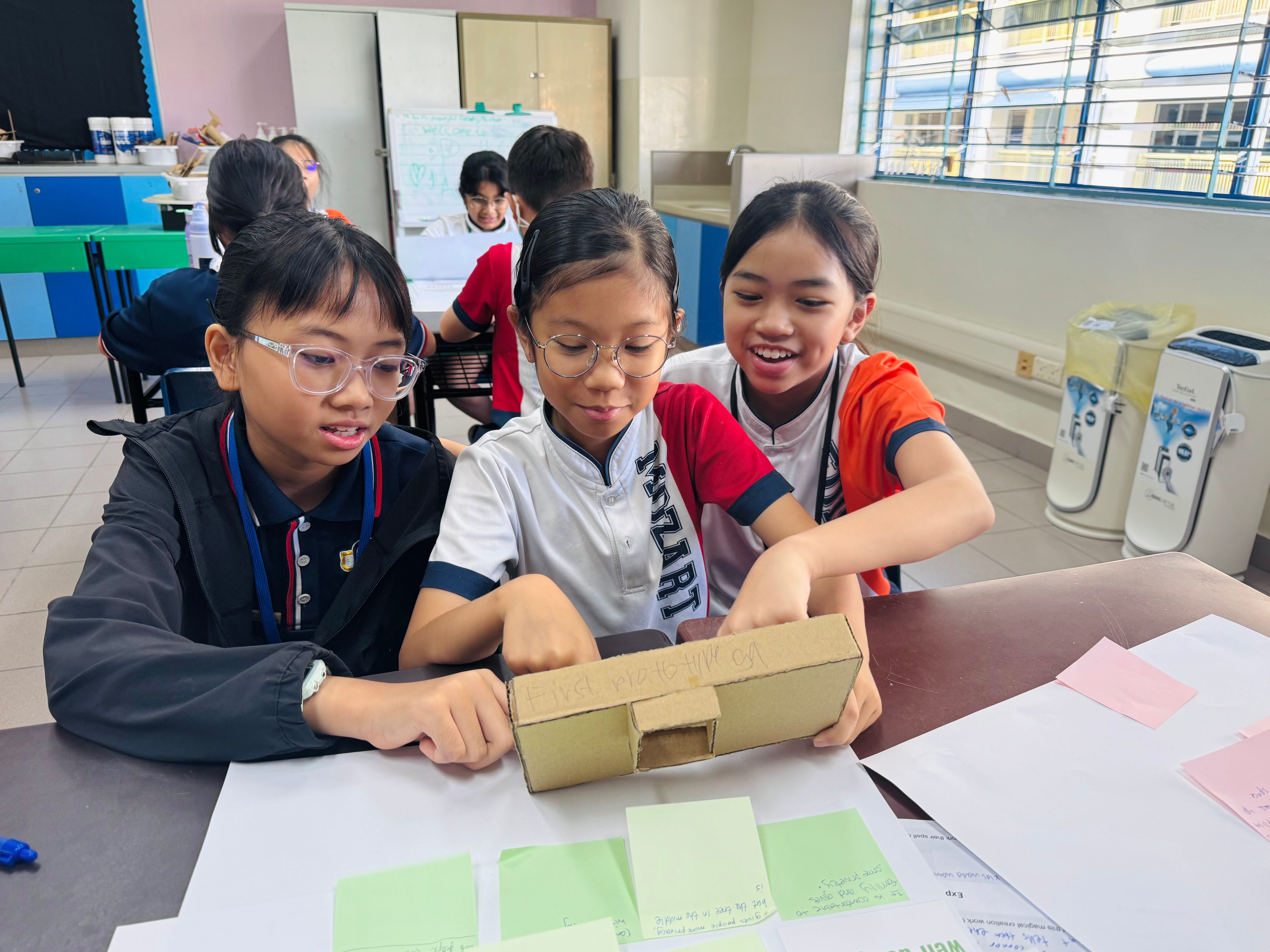 Three young girls in school uniforms collaborate on a cardboard model project at a classroom desk.