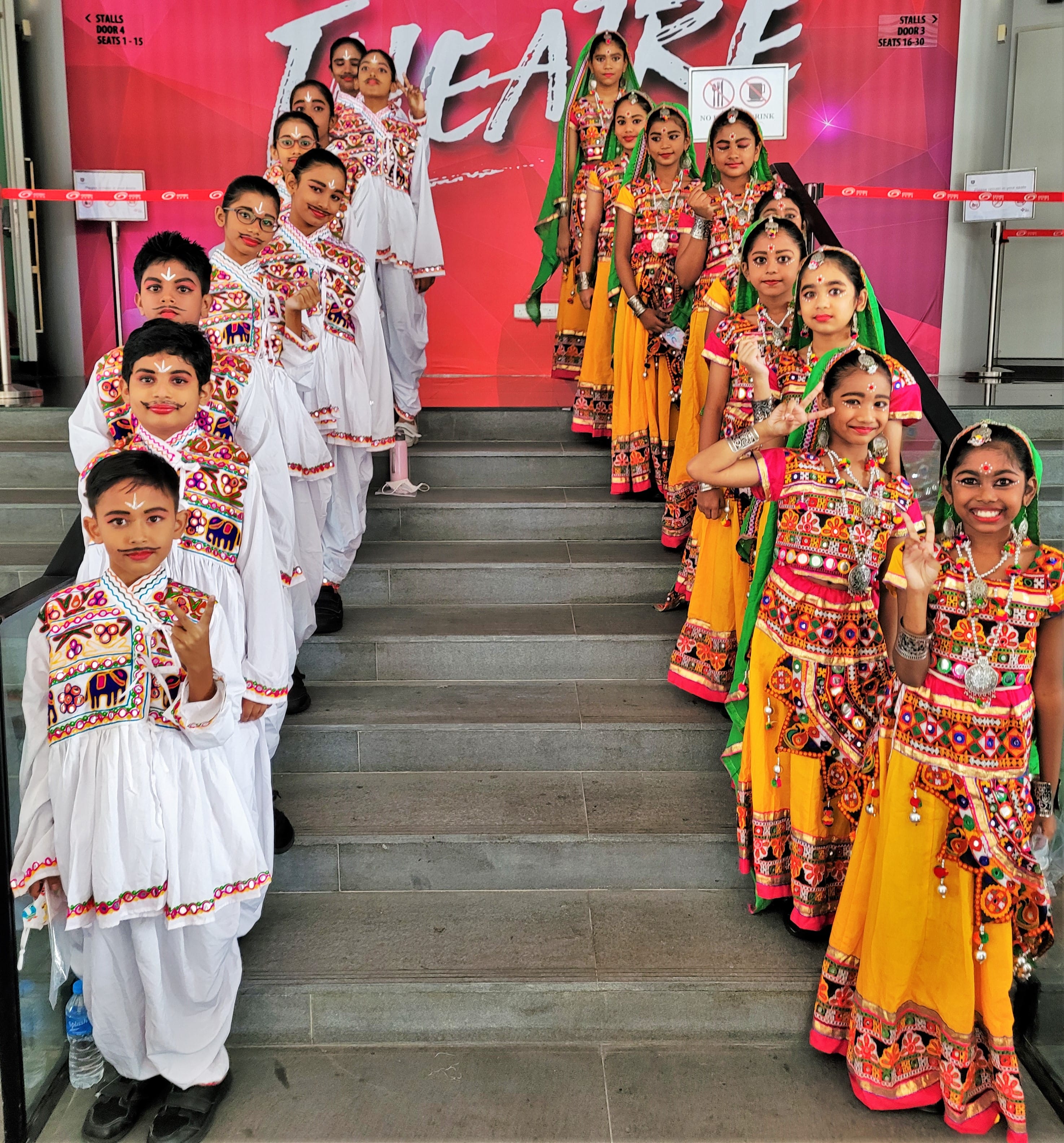 Children in colorful traditional outfits line both sides of steps, posing with hand signs before a red backdrop.