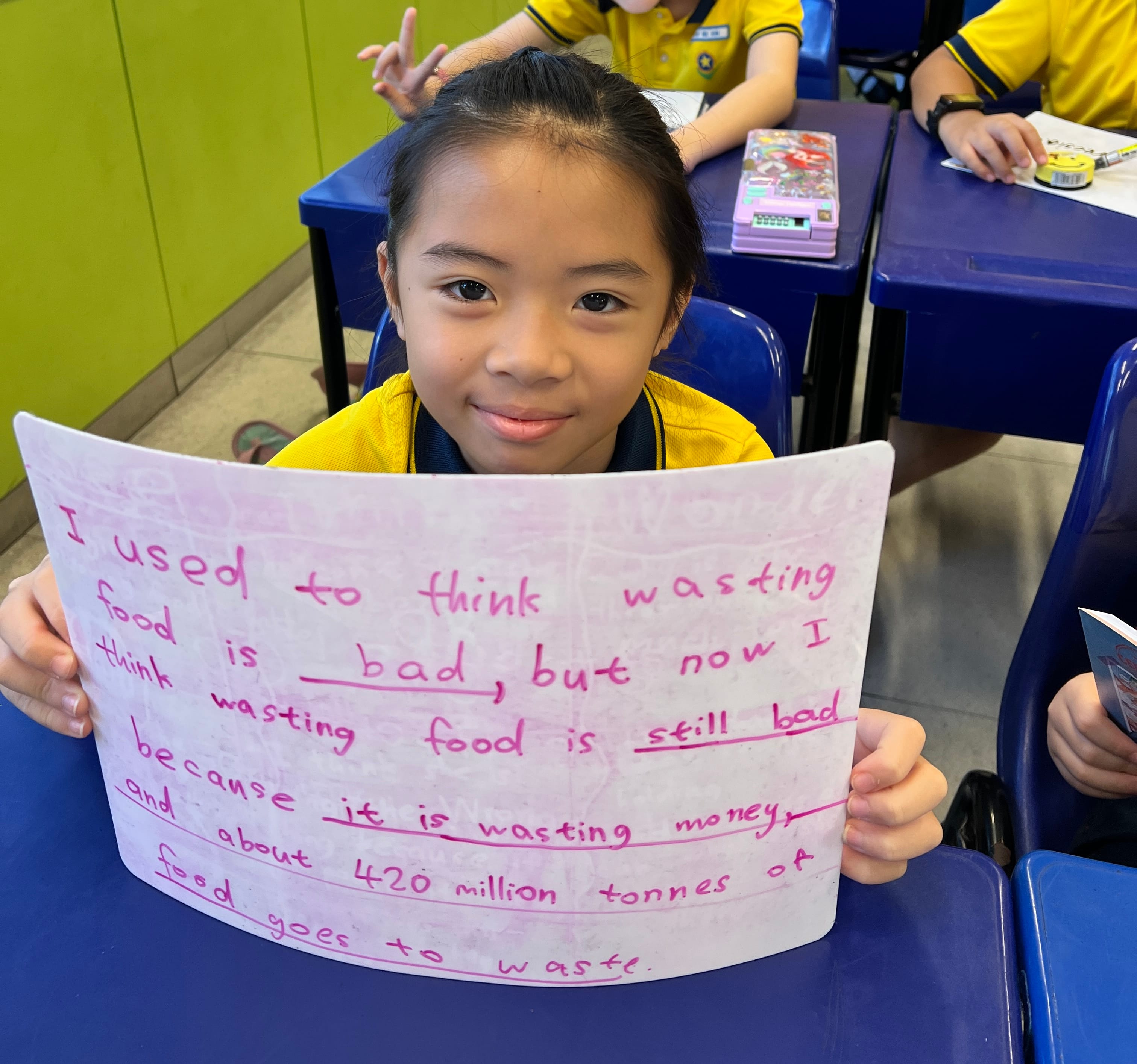 A smiling girl in a yellow uniform holds a handwritten poster about food waste in a classroom.