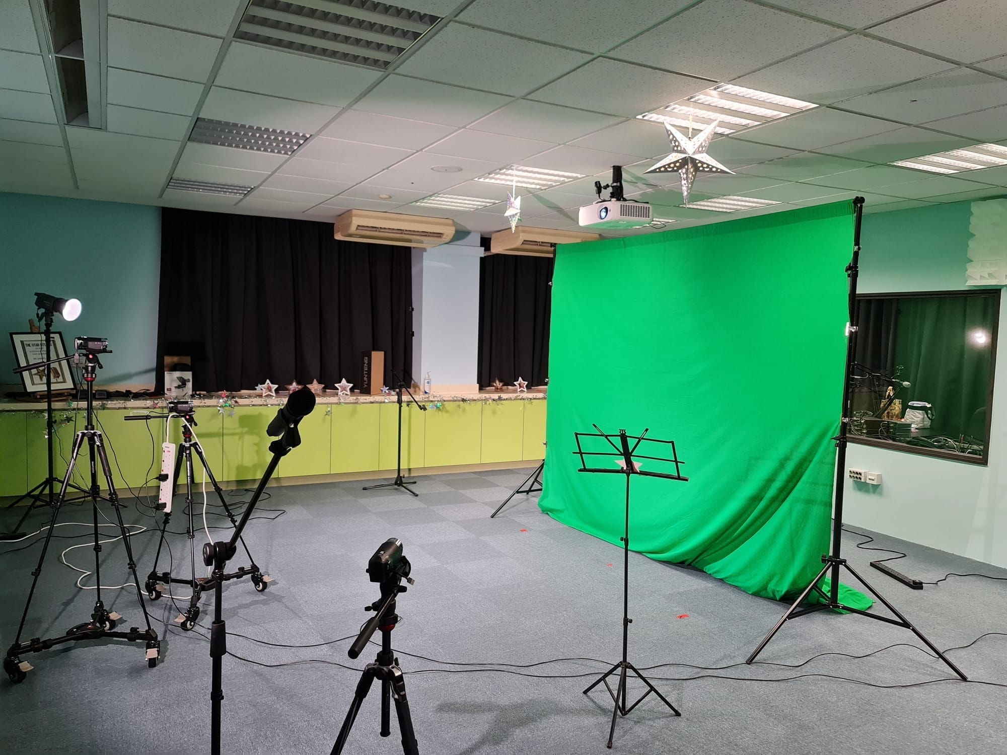 A video production setup with a large green screen, cameras on tripods, lights, and a music stand in a room.