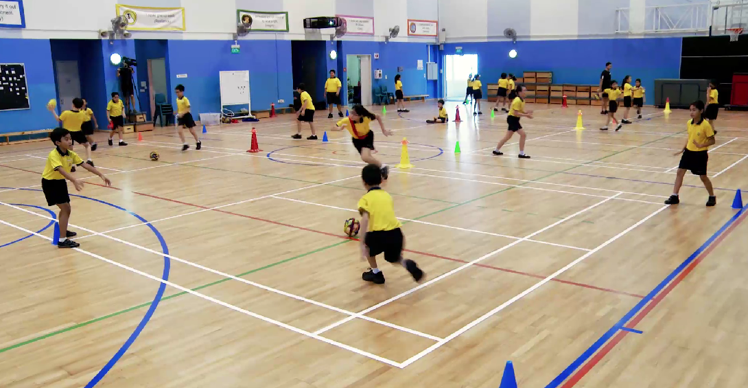 Children in yellow shirts dribble and play with balls in an indoor gymnasium with colored cones on the floor.