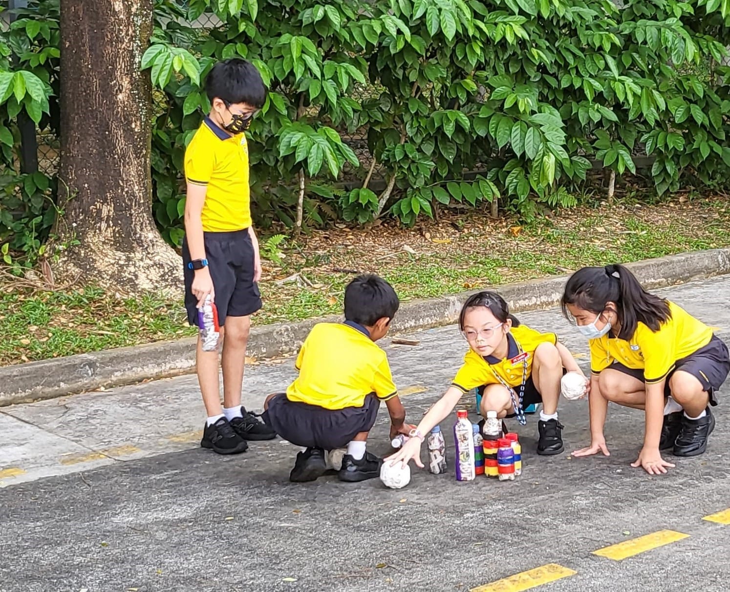 Four children in yellow school uniforms play with bottles and balls on a paved outdoor area near a tree.