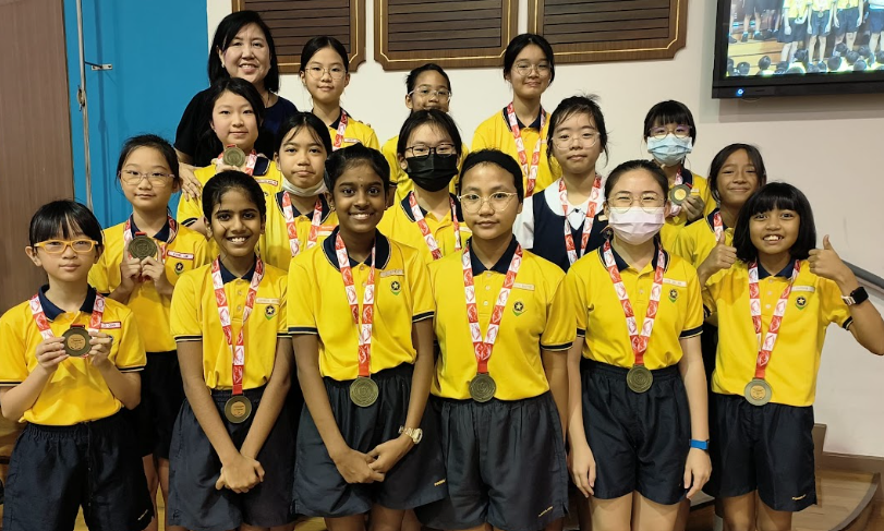 Group of young girls in yellow school uniforms proudly displaying medals, posing with an adult woman.