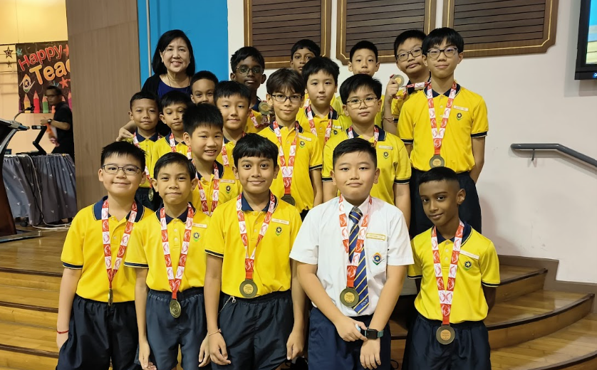 Group of young boys in yellow uniforms wearing medals, posing with a female teacher indoors.