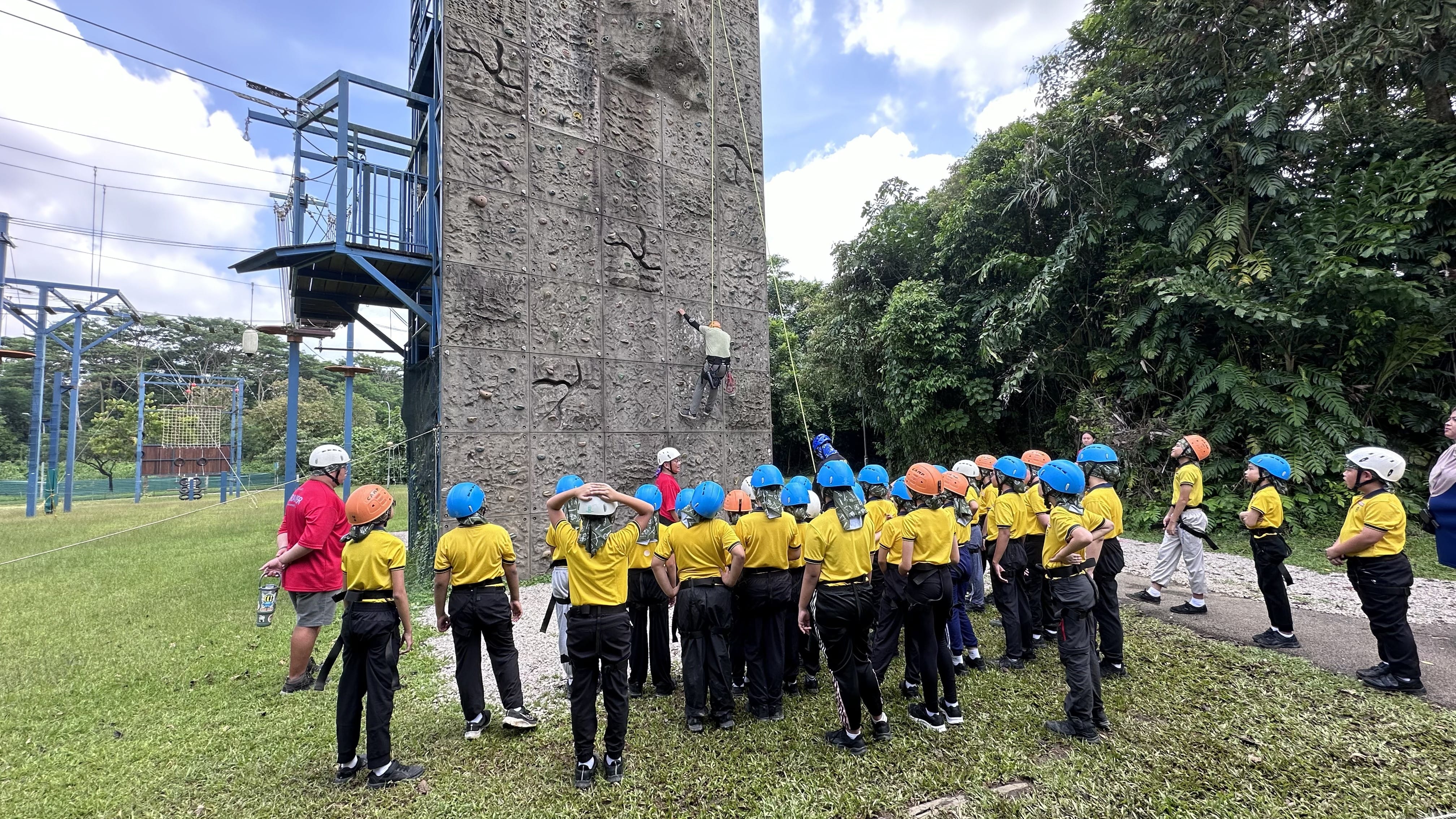 Students in yellow shirts and helmets watch a person climbing a tall outdoor rock wall near trees.