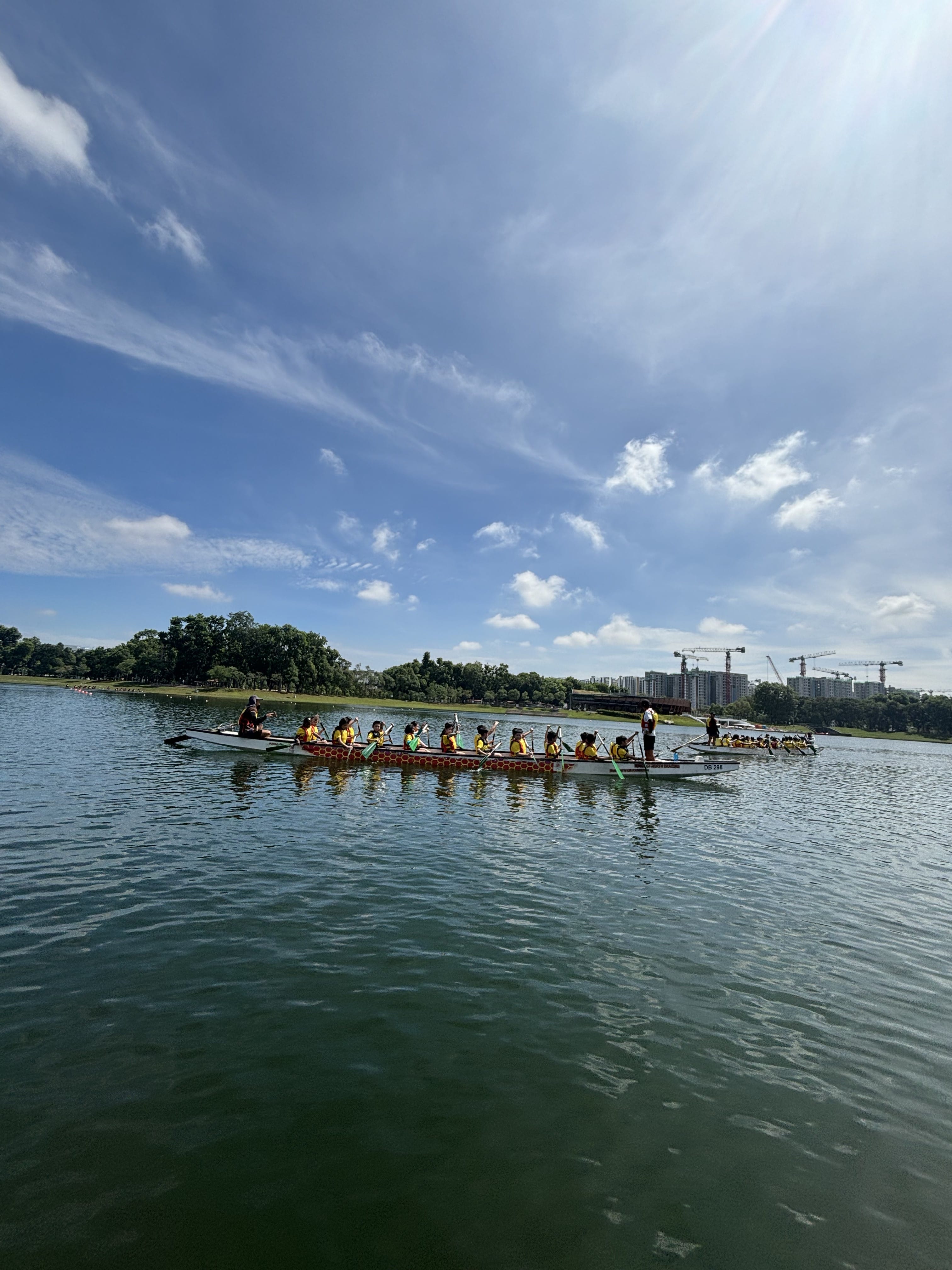 A dragon boat with paddlers in yellow vests rows across a calm lake under a bright blue sky with scattered clouds.