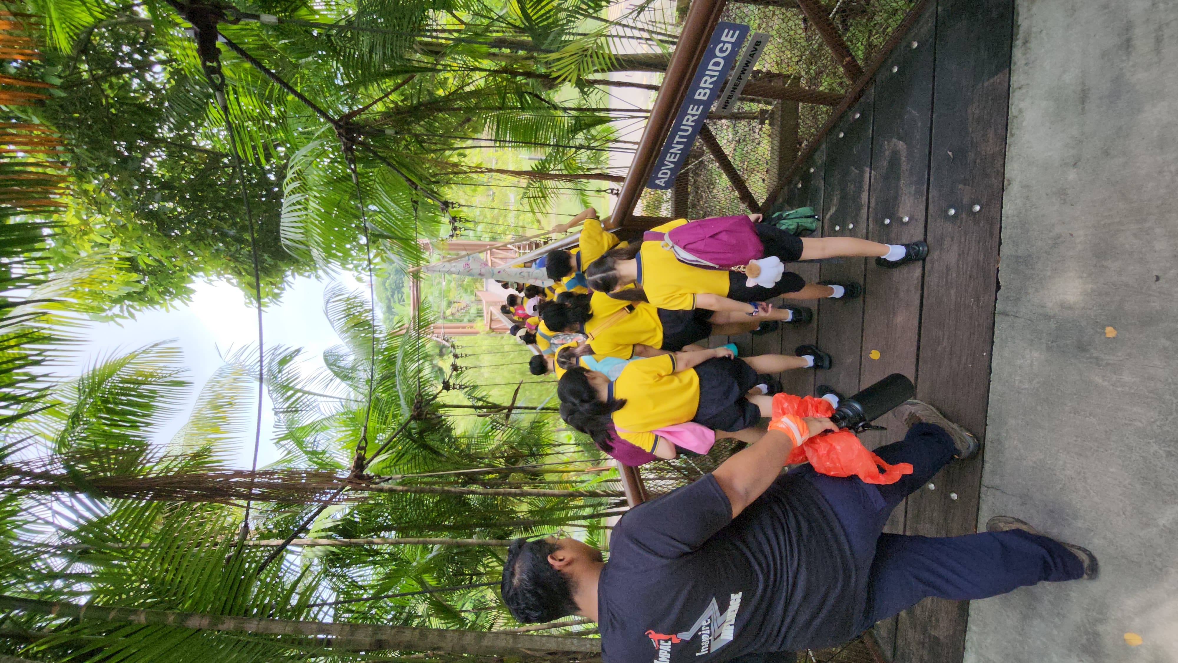 Students in yellow shirts cross a wooden suspension bridge labeled Adventure Bridge surrounded by tropical trees.