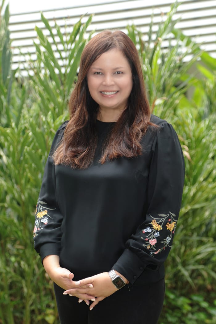 Woman with long brown hair smiling, wearing black top with floral embroidered sleeves, standing before green plants.
