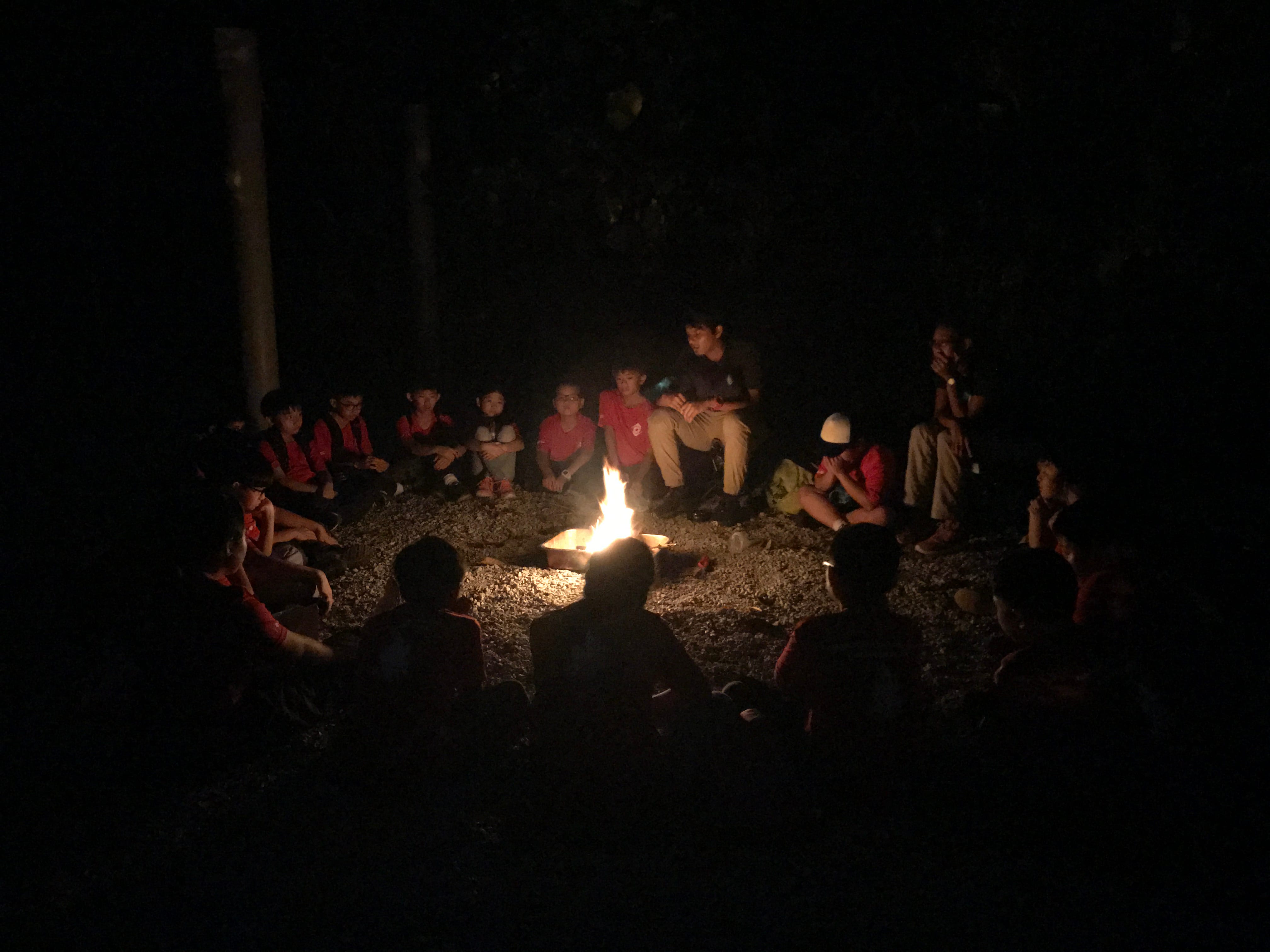A group of children and adults sit in a circle around a small campfire at night outdoors.