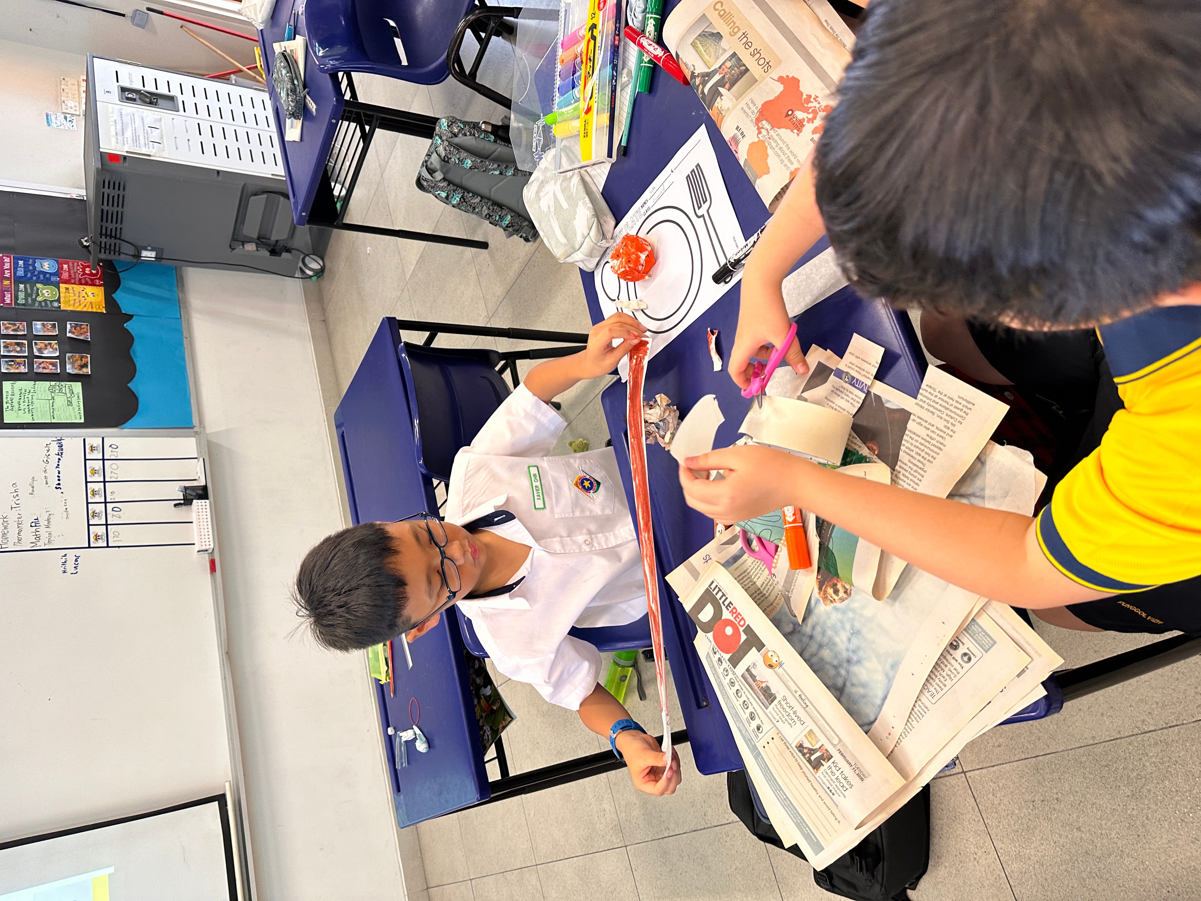 Two school children work on a craft project at a blue desk covered with newspaper, scissors, and markers.