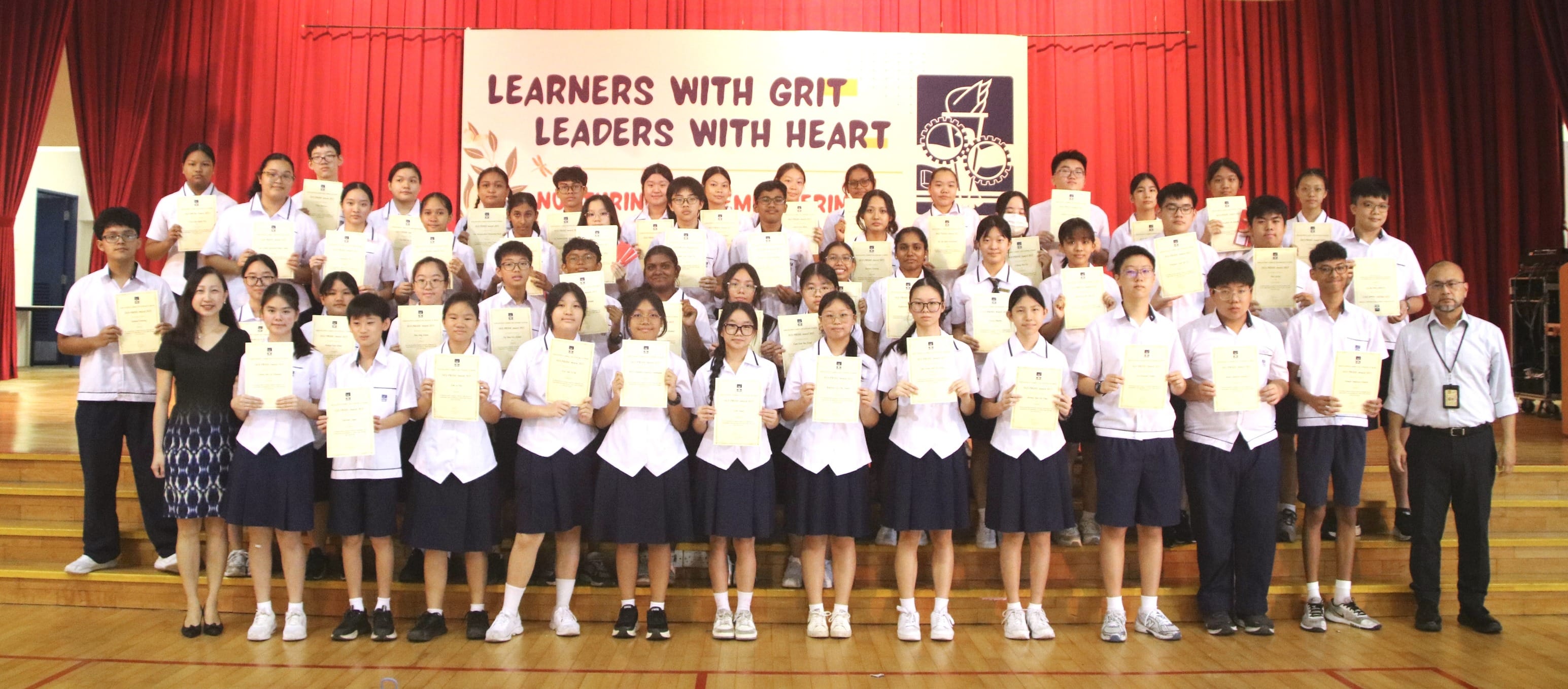 Large group of school students in uniforms holding certificates on a stage with a banner reading Learners With Grit Leaders With Heart.