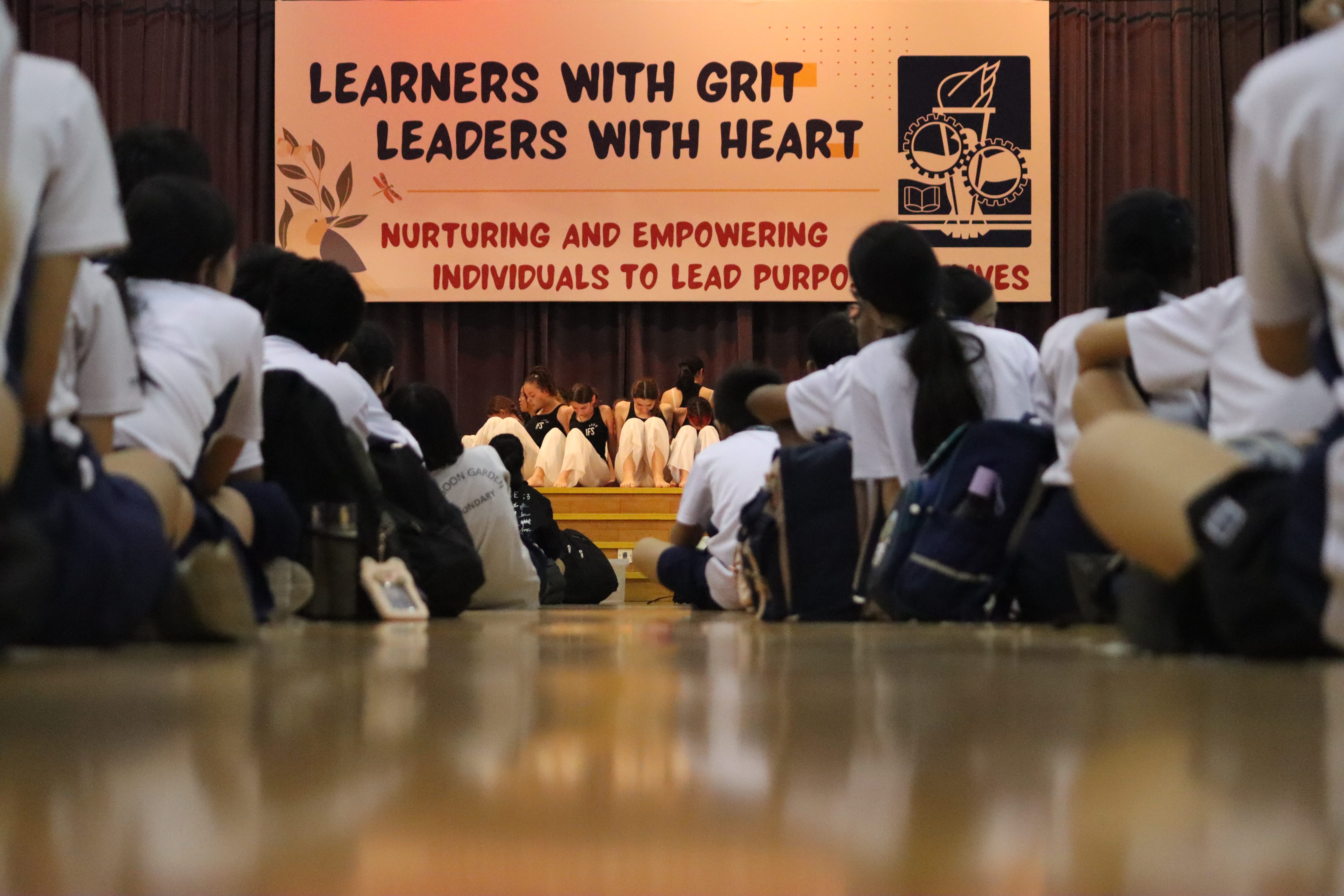 Students sit on a gym floor facing a stage under a banner reading Learners with Grit, Leaders with Heart