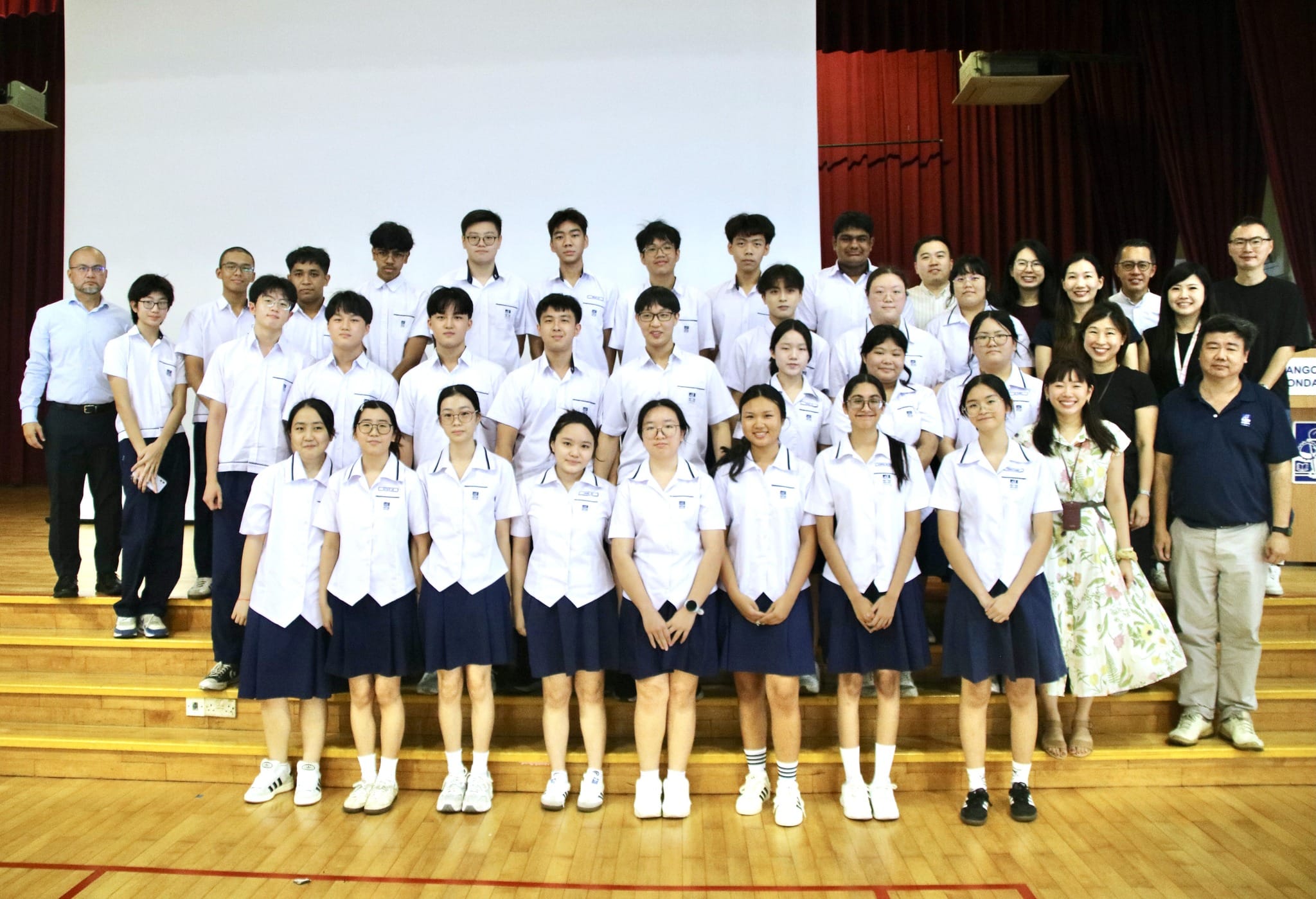 A large group of students in white and navy uniforms pose with adults on a school stage in front of a white screen.