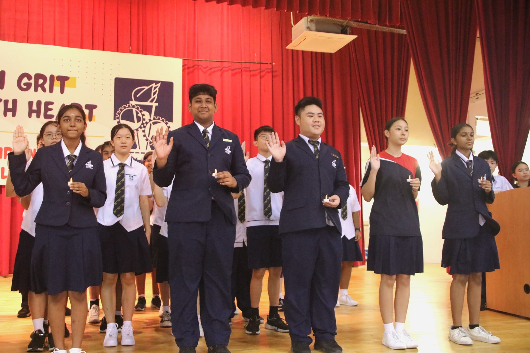 Students in school uniforms raise their right hands during a pledge ceremony on a stage with red curtains.